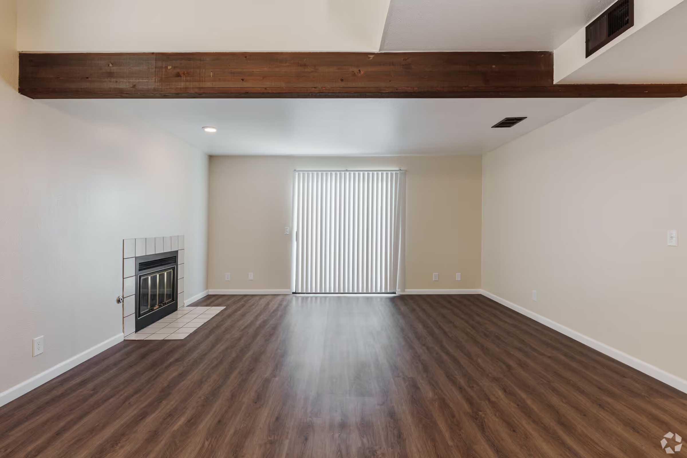 Spacious room featuring a central fireplace with a wooden beam overhead, light-colored walls, and large windows partially covered by vertical blinds. The floor is finished with a wood-like laminate, creating a modern, inviting atmosphere.