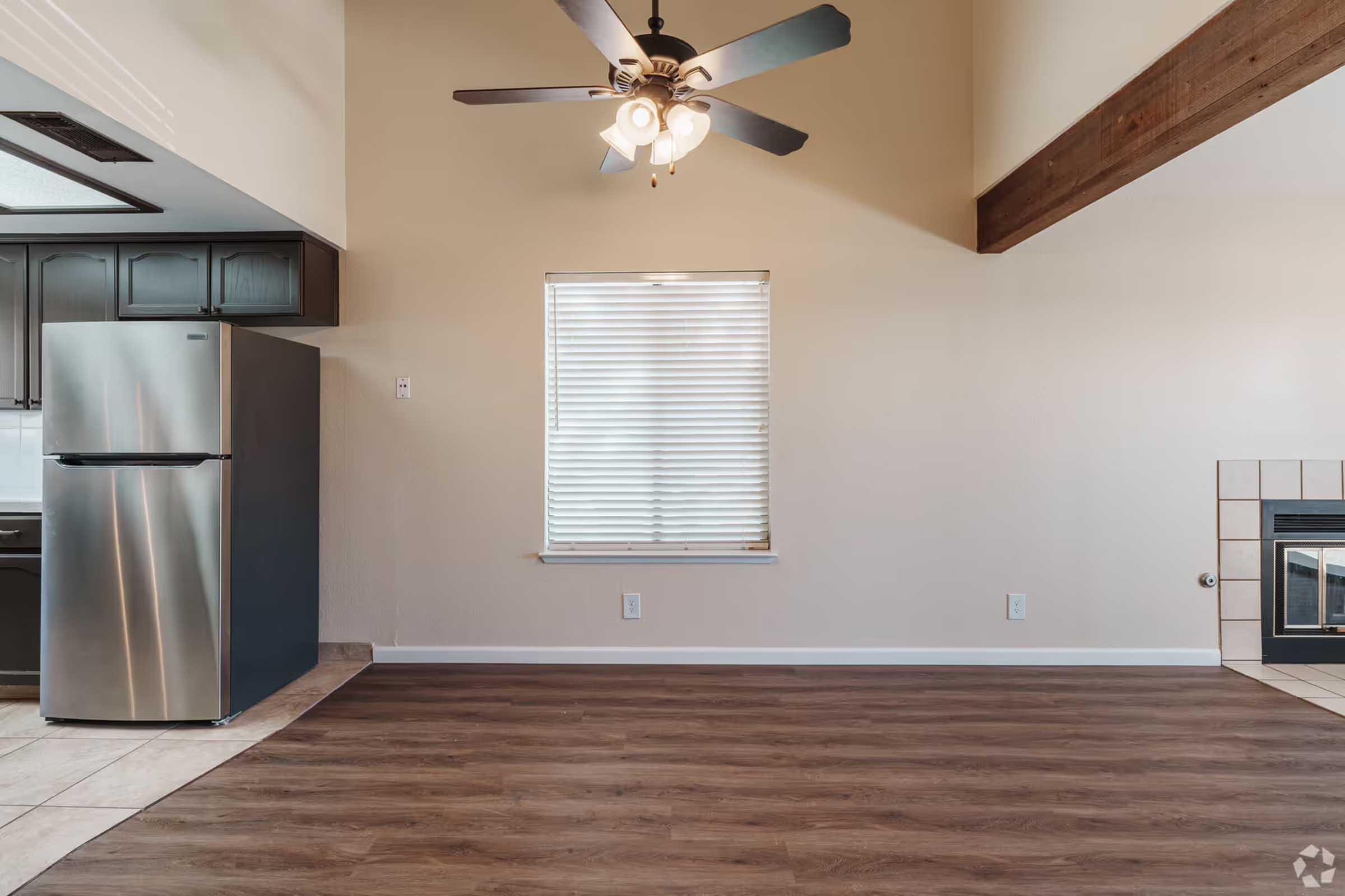 Interior view of a spacious living area featuring a ceiling fan with light fixtures, a large window with blinds, and a sleek stainless steel refrigerator in the adjacent kitchen. The space has hardwood-like flooring and a cozy fireplace visible on the right. Soft, neutral wall colors complement the overall design.