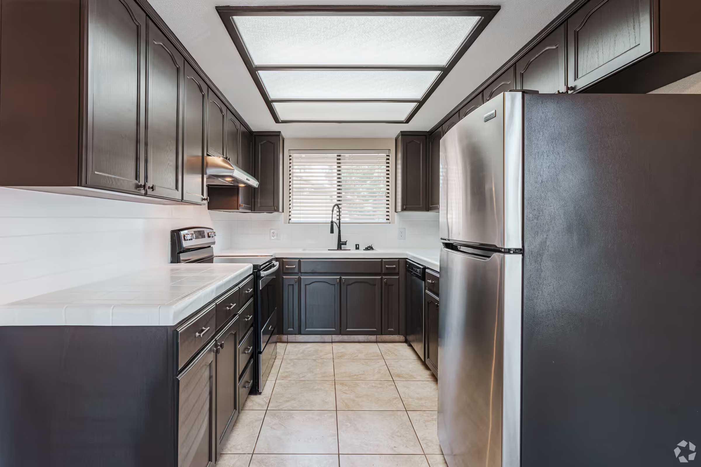 Modern kitchen featuring dark cabinetry, stainless steel appliances, and a light-colored countertop. The room has a tile floor and a window with blinds, allowing natural light to enter. A spacious layout includes a stove, oven, refrigerator, and sink, creating a functional cooking space.