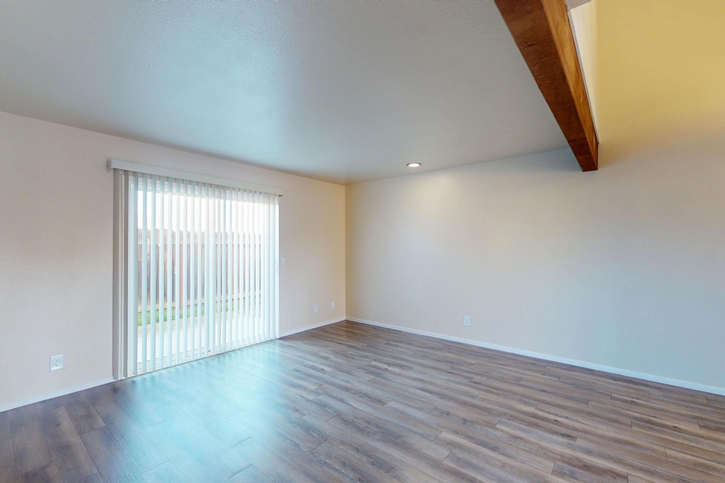 A spacious, empty room with light-colored walls and wood-style flooring. Large sliding glass doors covered with vertical blinds allow natural light to enter, while a wooden beam runs along the ceiling, adding architectural interest. The space is ready for furnishing and decoration.