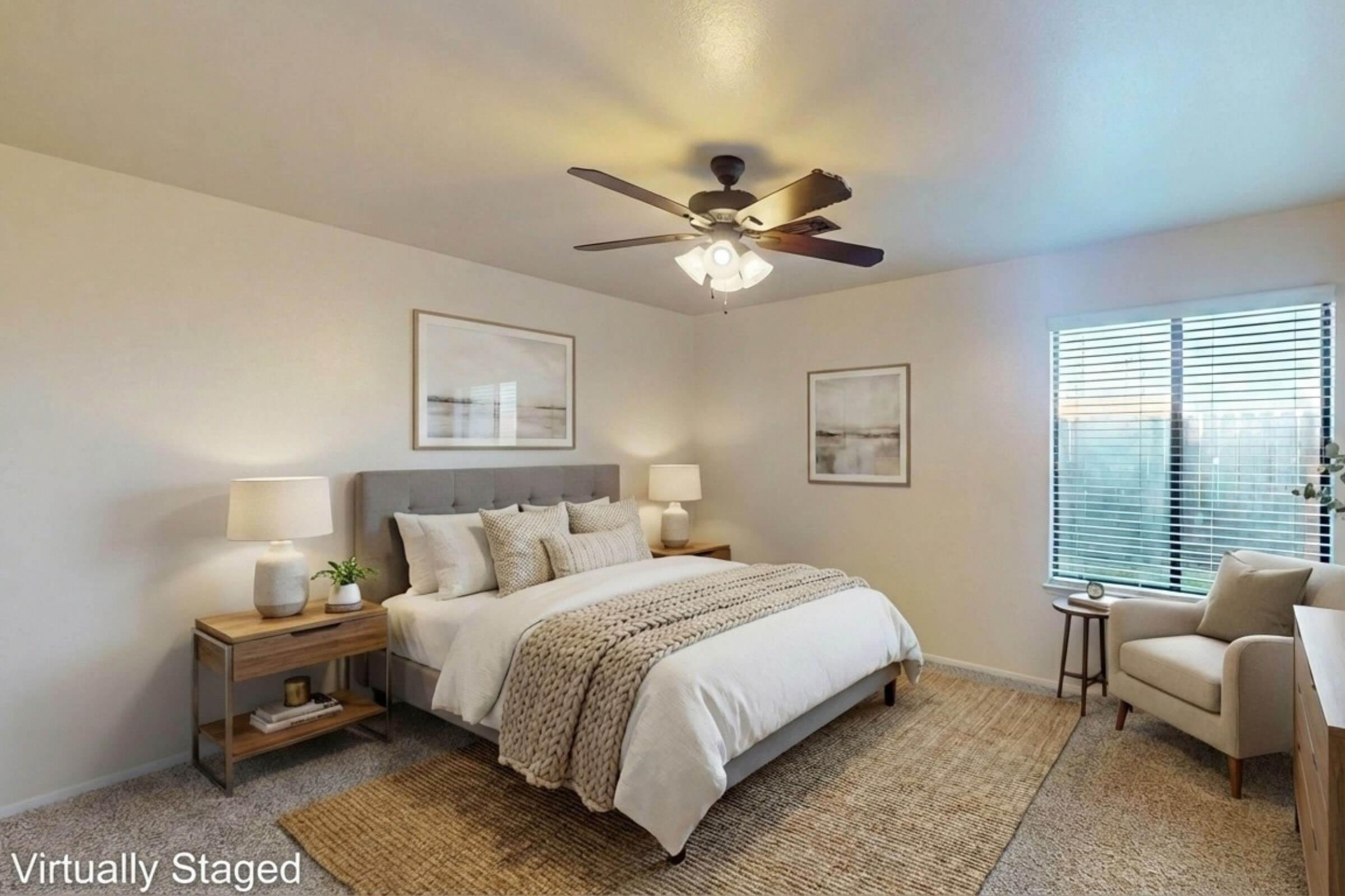 A bright, modern bedroom featuring a queen-sized bed with light bedding and decorative pillows. There are two bedside tables with lamps, a cozy armchair by the window, a ceiling fan, and artwork on the walls. Natural light streams in through the window, illuminating the space elegantly.