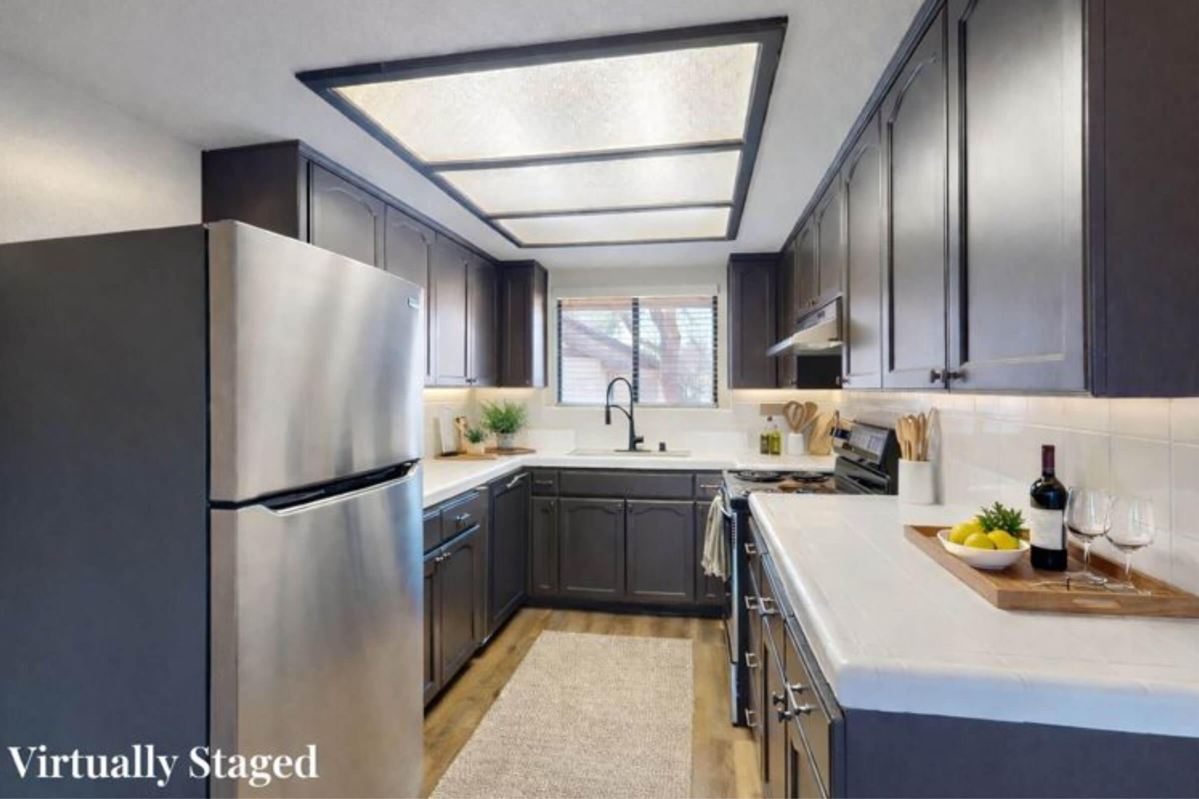 A modern kitchen featuring dark cabinetry, a stainless steel refrigerator, and white countertops. The space is well-lit with a ceiling light, showcasing a sink with a black faucet, a stove, and decorative elements like a fruit bowl and wine glasses on a tray. A light-colored rug adds warmth to the floor.