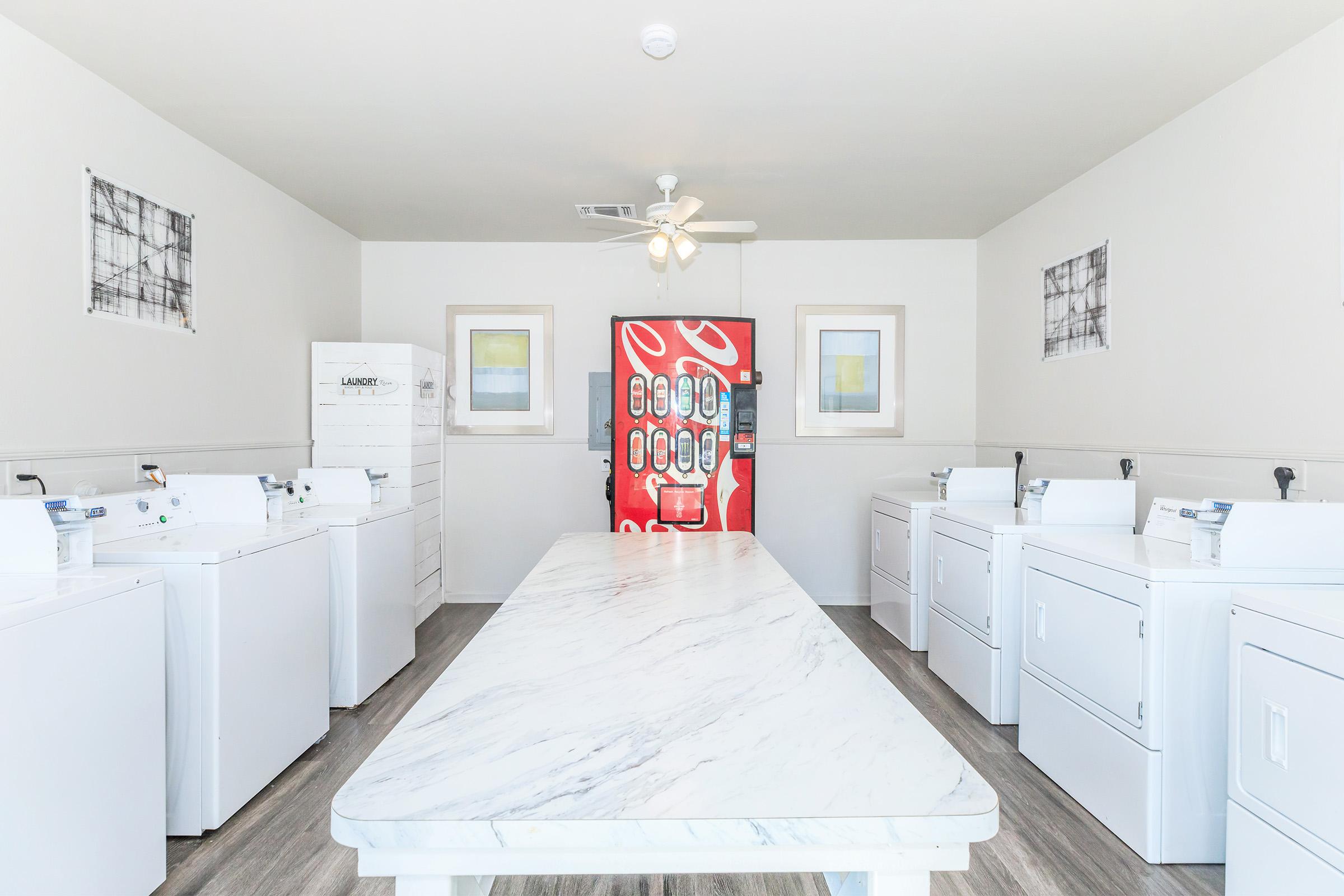 A bright and clean laundry room featuring several white washing machines and dryers arranged in rows. The room is illuminated by a ceiling fan and has light-colored walls adorned with abstract art. In the background, there's a bright red vending machine with various snacks and drinks.