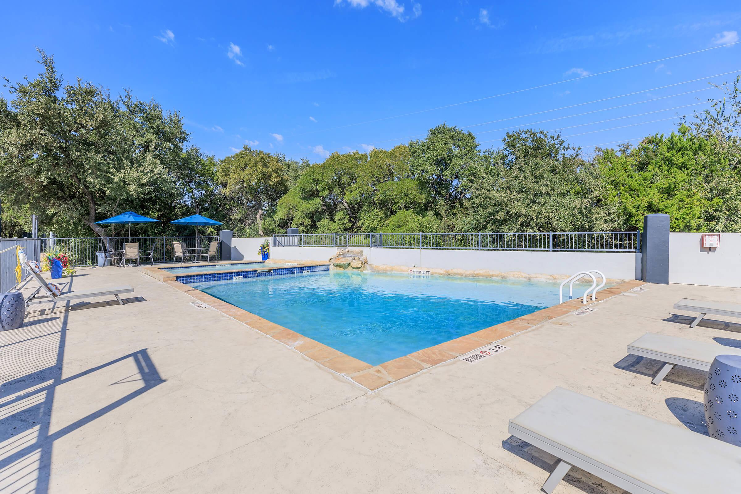 A well-maintained outdoor swimming pool surrounded by a spacious deck, featuring lounge chairs and shaded seating areas. Lush green trees are visible in the background under a clear blue sky, creating a serene atmosphere for relaxation and leisure.