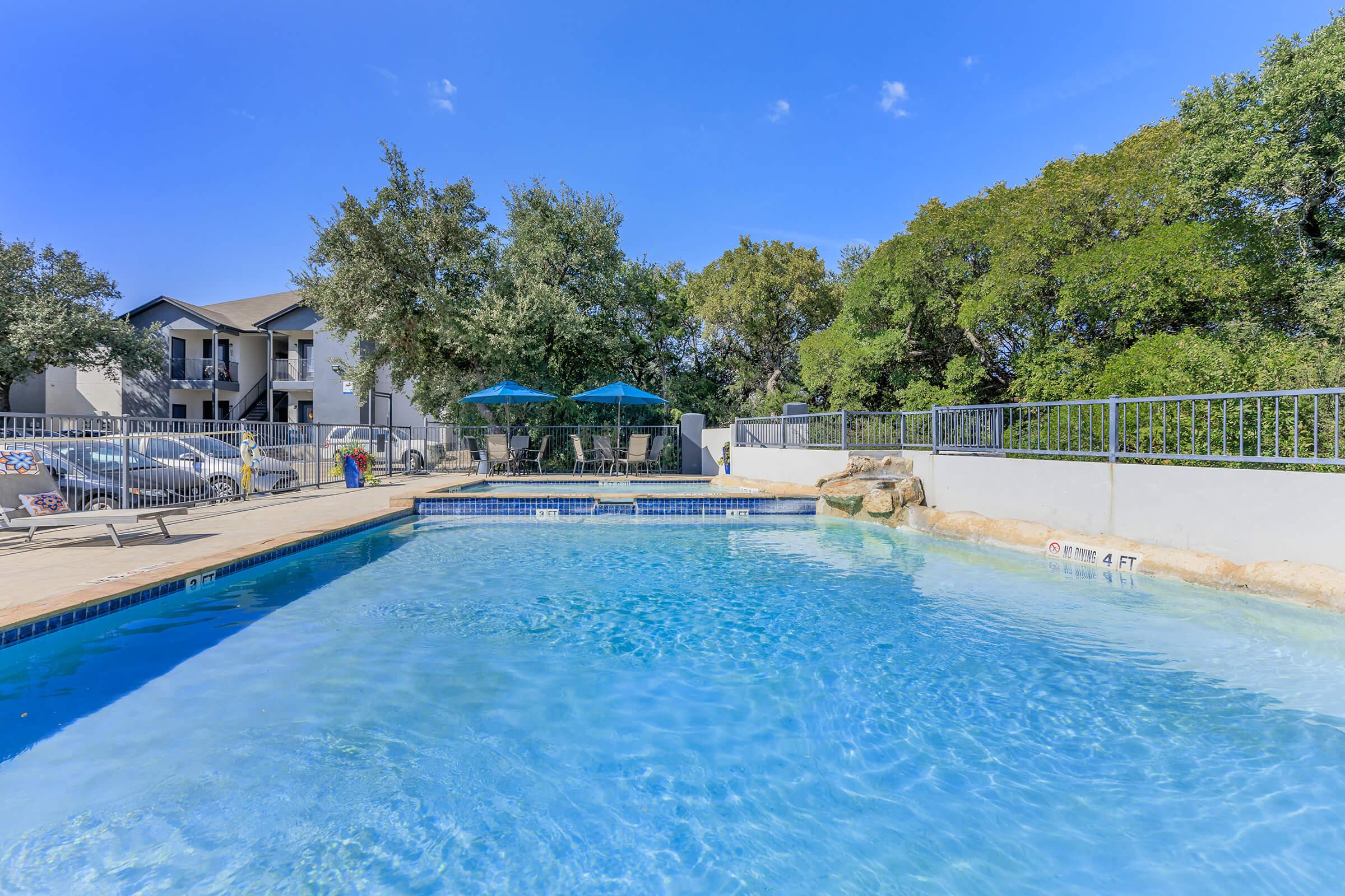 A clear blue swimming pool surrounded by a deck with lounge chairs, shaded by umbrellas. In the background, there is a row of buildings and trees, creating a serene outdoor setting. The pool features a shallow area marked "4 FT." and is under a clear blue sky.