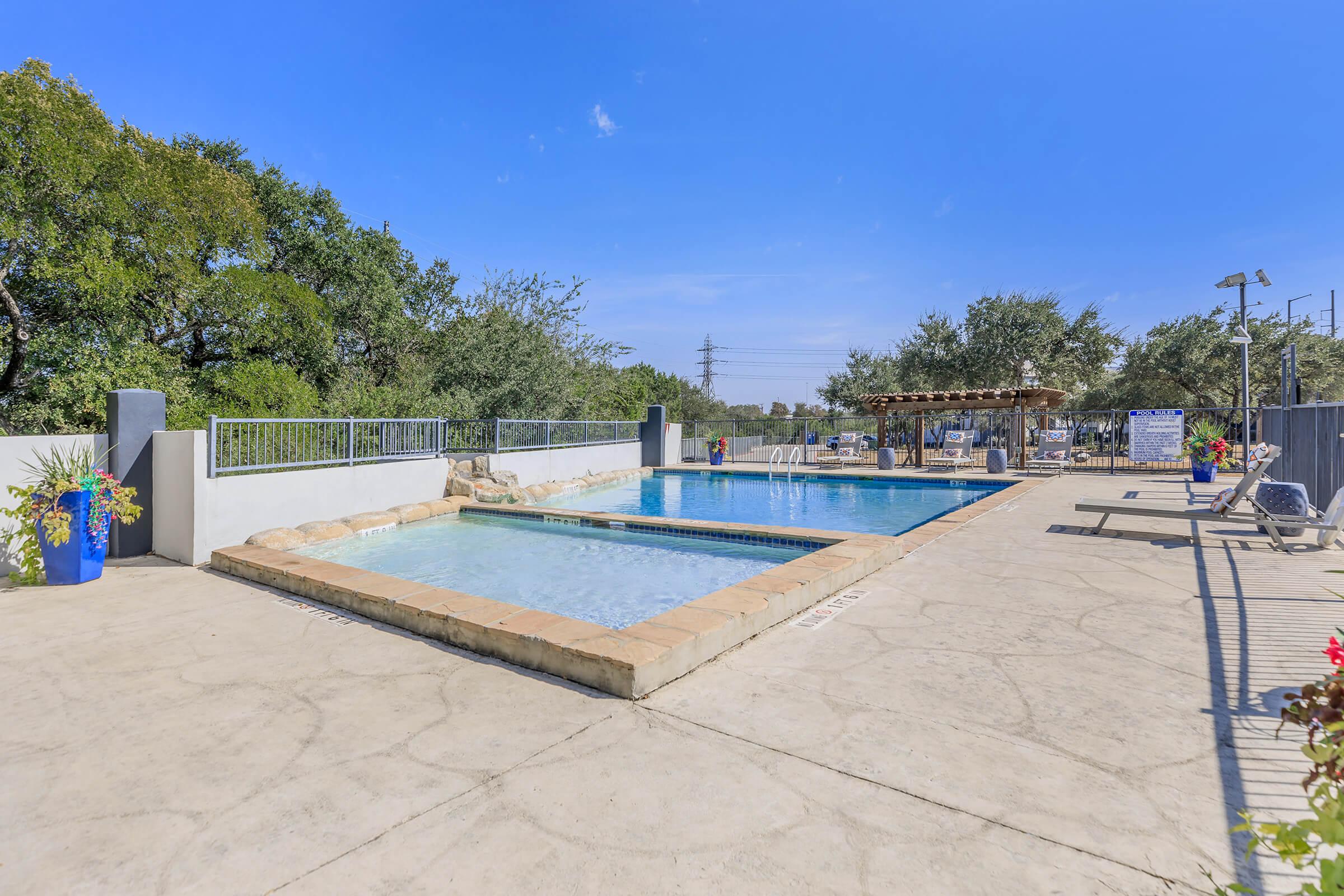 A clear blue sky over a swimming pool area featuring two pools: one shallow and one deeper, surrounded by paved flooring. There are lounge chairs set up for relaxation, lush greenery nearby, and a shaded pergola space with seating. Lighting fixtures are visible, indicating a well-maintained outdoor recreational area.