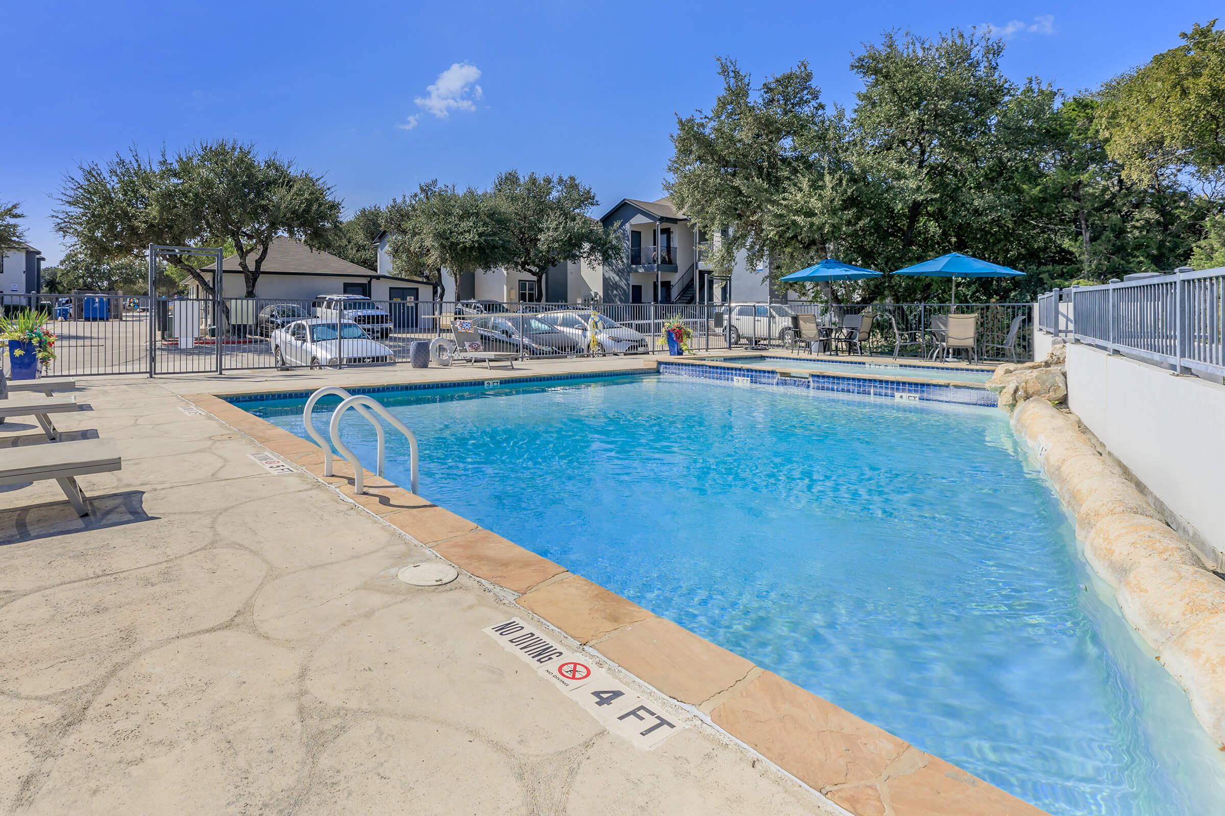 A clear swimming pool with a shallow end marked at 4 feet, surrounded by a concrete deck. There are lounge chairs and umbrellas nearby, with trees and residential buildings in the background. The sky is blue with a few clouds.