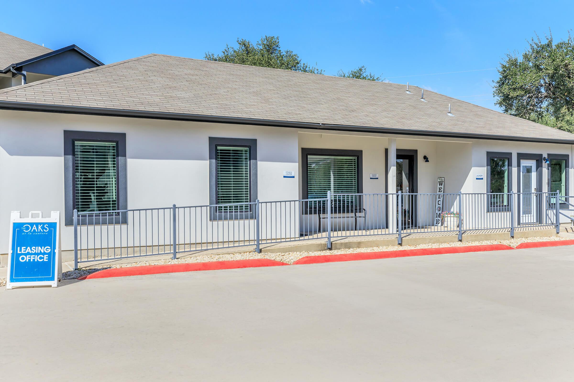 A modern leasing office building with a light-colored exterior and multiple windows, located on a paved area. There is a sign in front indicating it is the "OAKS LEASING OFFICE." The surrounding area features greenery and a clear blue sky.