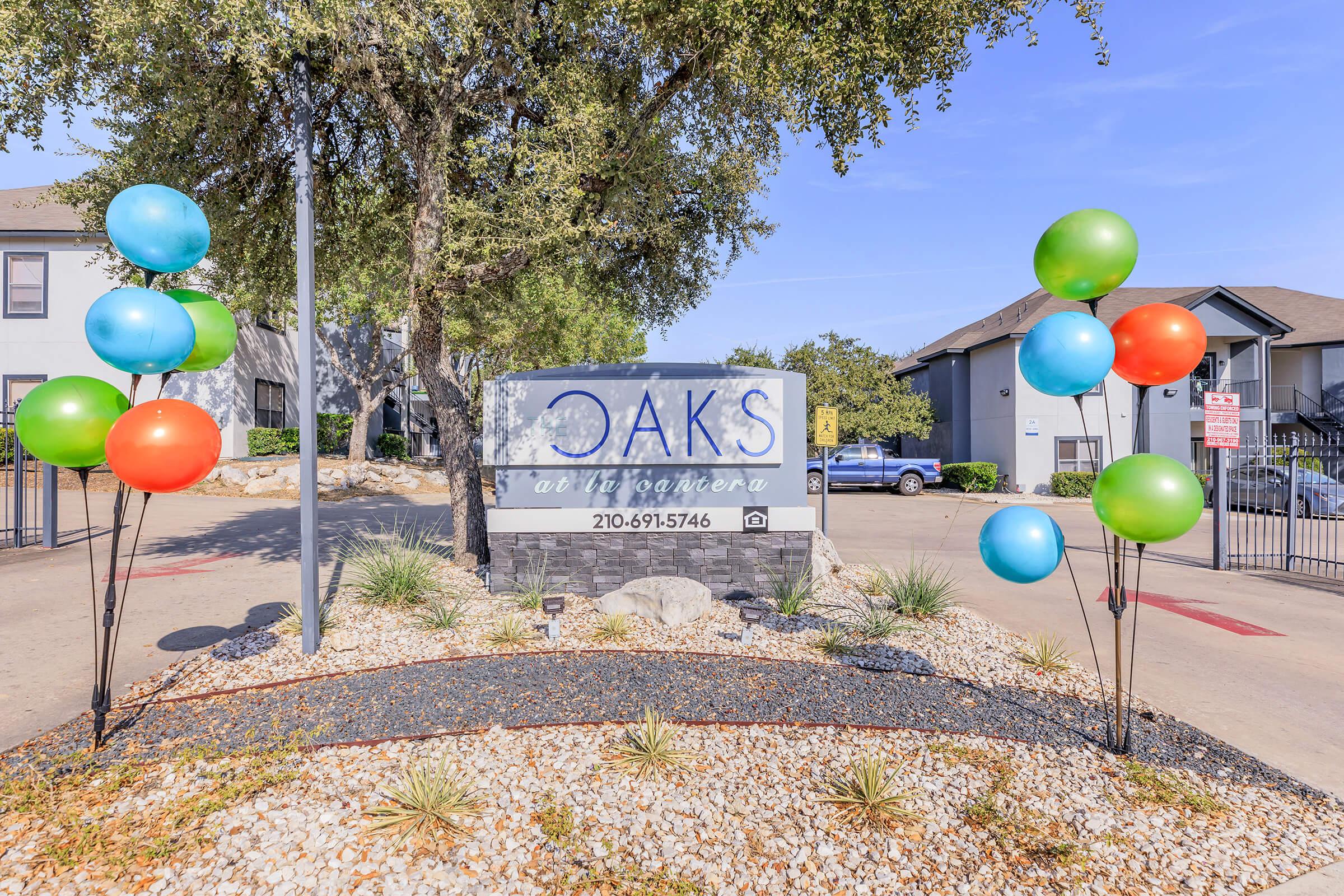 A colorful entrance sign for "Oaks at the Linconera" featuring green and orange balloons on either side. The sign is surrounded by gravel, plants, and small rocks, set against a clear blue sky and nearby apartment buildings in the background.