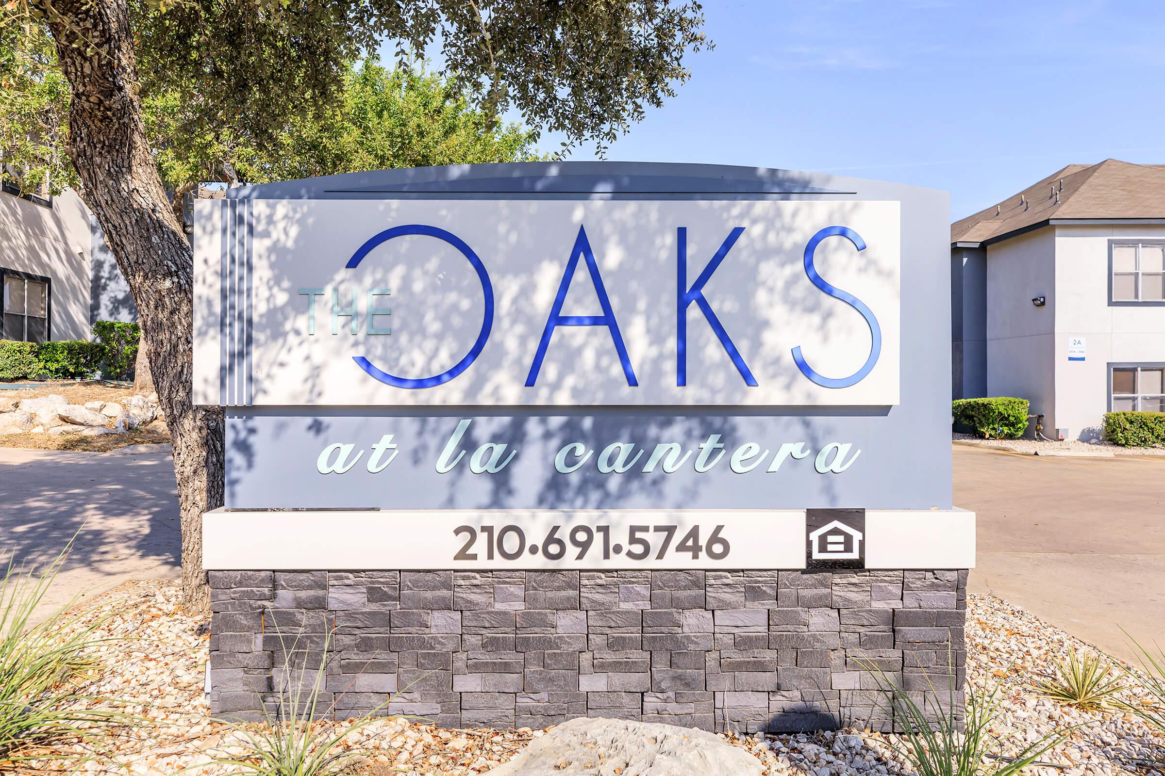 A sign for "The Oaks at La Cantera" featuring the name prominently displayed in blue letters. The sign includes a phone number, 210-691-5746, and is surrounded by landscaping with rocks and shrubs. The background shows a building and trees.