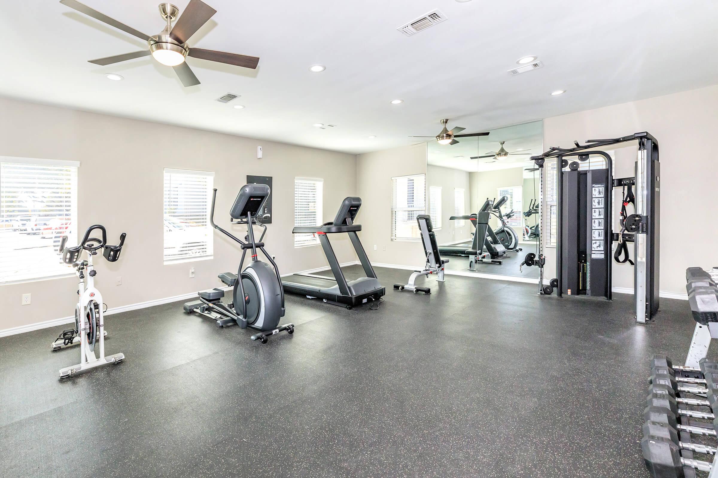 A modern gym space featuring various fitness equipment including treadmills, exercise bikes, and weight machines. The room has a clean design with large windows allowing natural light, ceiling fans, and rubber flooring for comfort.