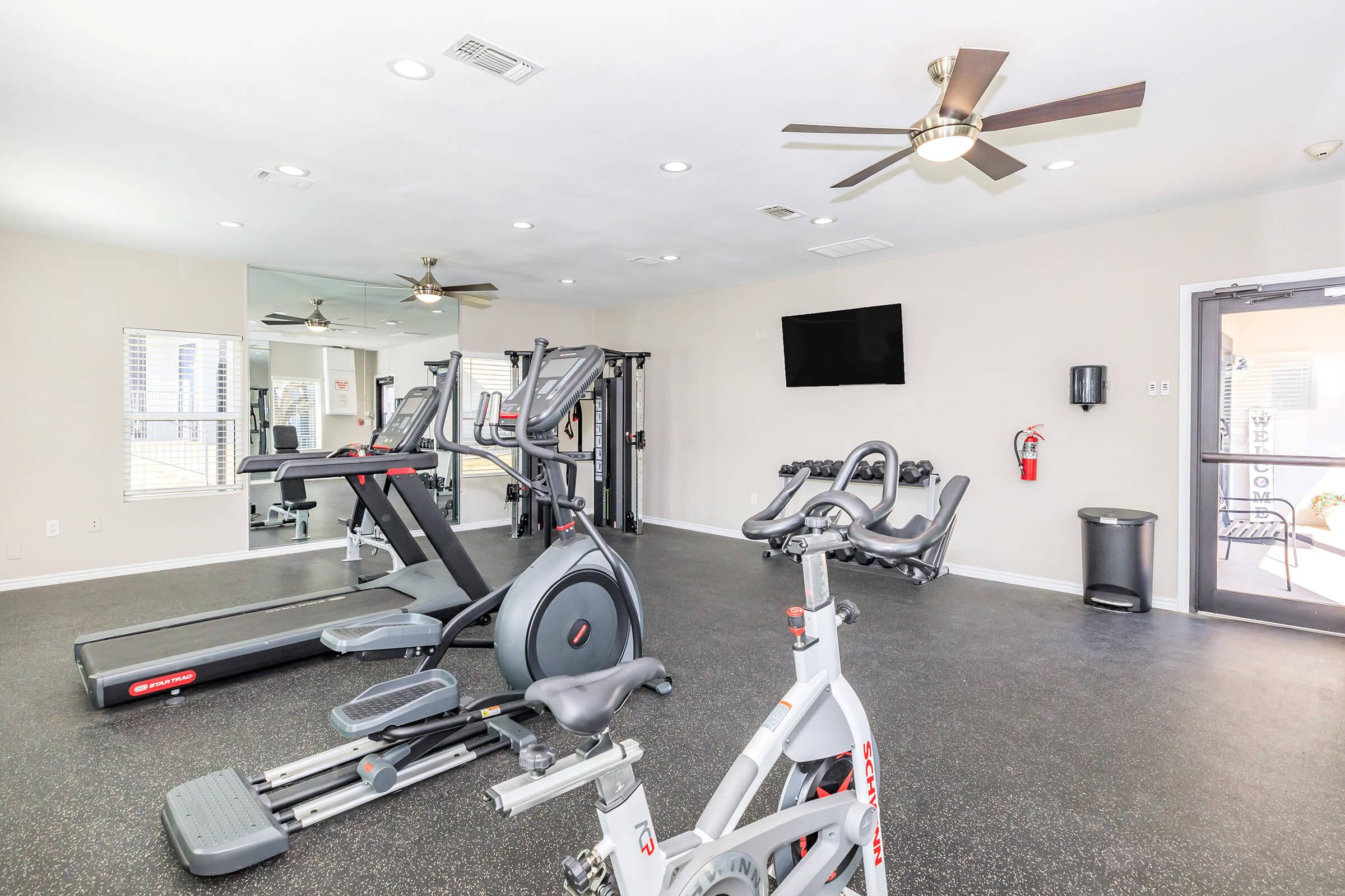 A spacious gym area featuring various exercise equipment including treadmills, elliptical machines, and stationary bikes. The room has ceiling fans, mirrors, a wall-mounted television, and is well-lit with natural light coming from windows. There's also a fire extinguisher and a trash can visible.