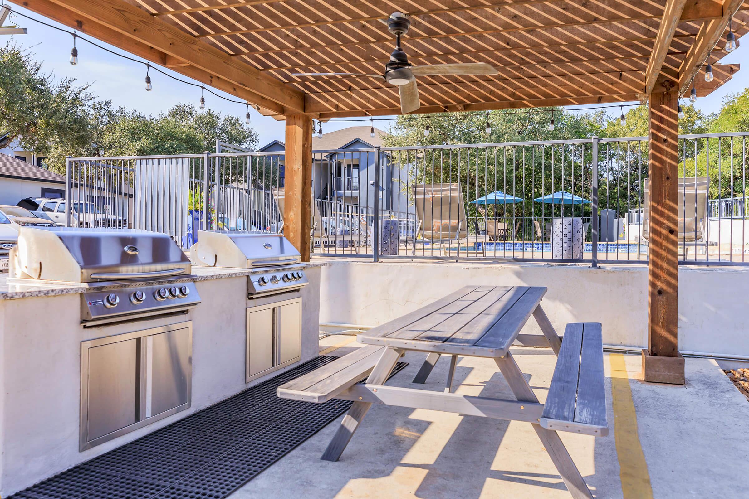 Outdoor grilling area featuring two stainless steel barbecue grills beneath a wooden pergola. A wooden picnic table is positioned nearby on a concrete surface. In the background, a fenced pool area with lounge chairs and trees can be seen, creating a relaxing social space.