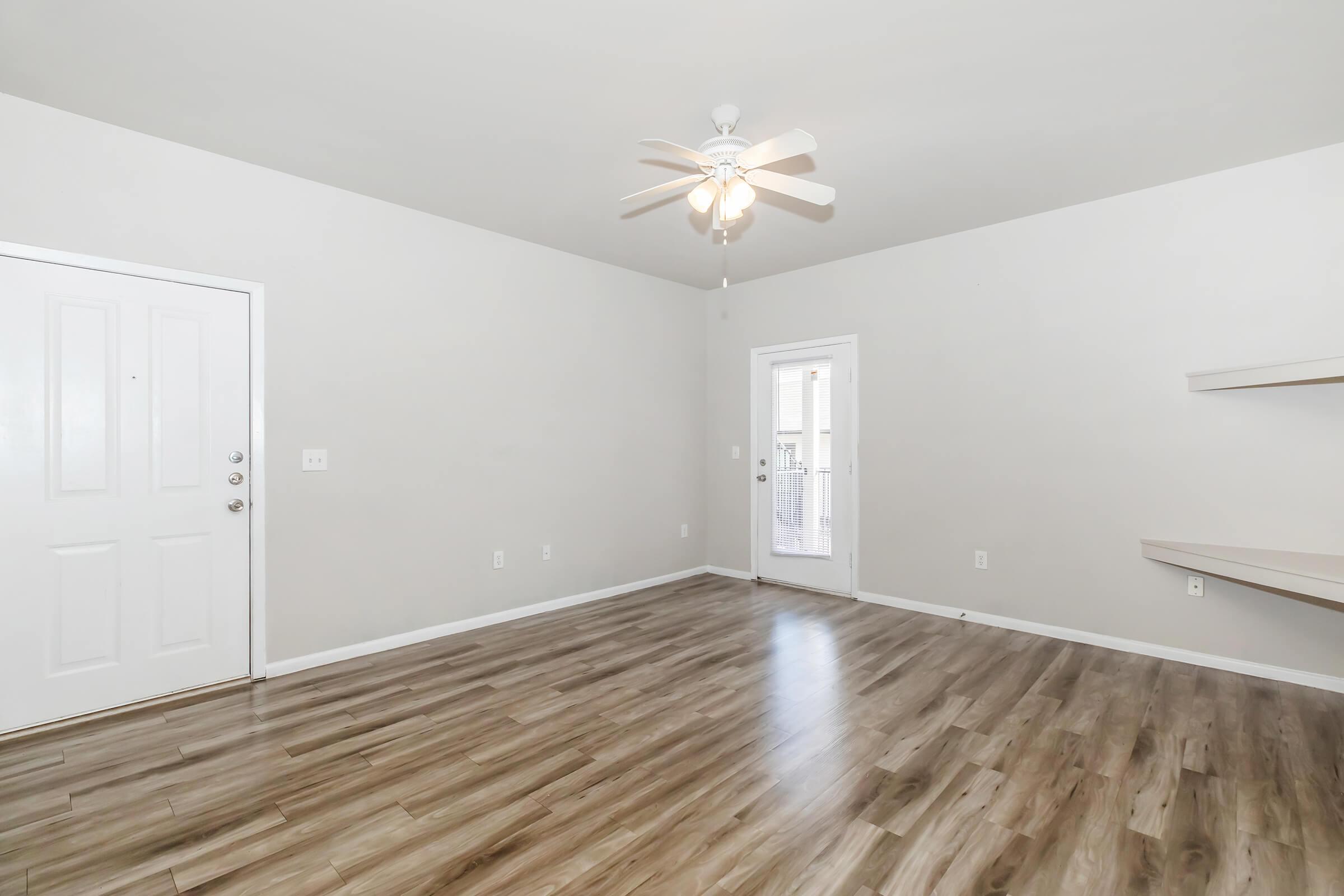 A spacious, empty living room featuring light gray walls, a ceiling fan, and laminate wood flooring. It includes a front door with a small window and a nearby sliding glass door leading to an outdoor area. A simple shelf is mounted on the wall beside the door.