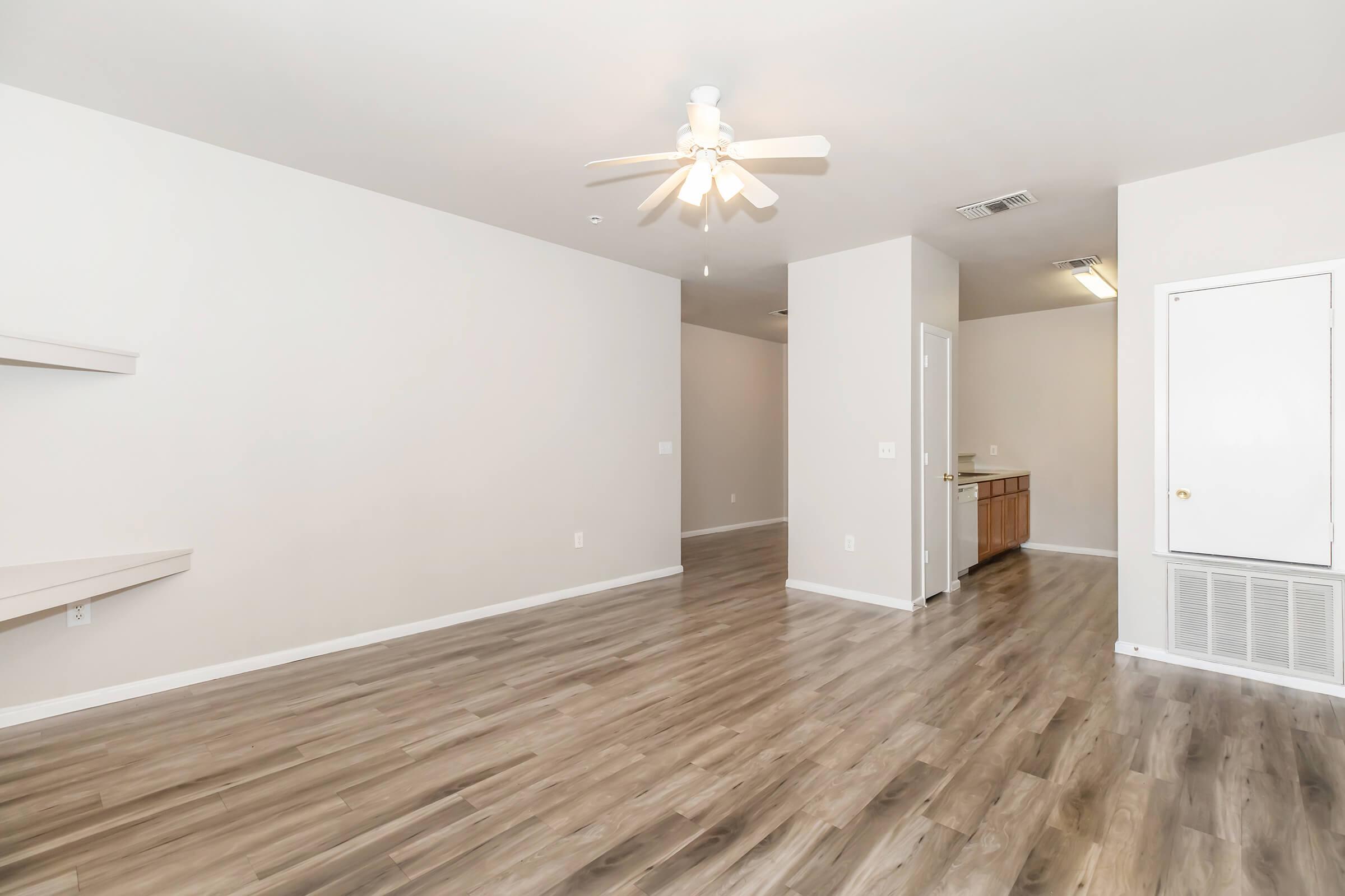 A spacious, neutral-colored living area featuring light wooden flooring, a ceiling fan, and simple wall shelving. In the background, there's an open kitchen with wooden cabinets. The overall design is modern and minimalistic, creating a bright and inviting atmosphere.