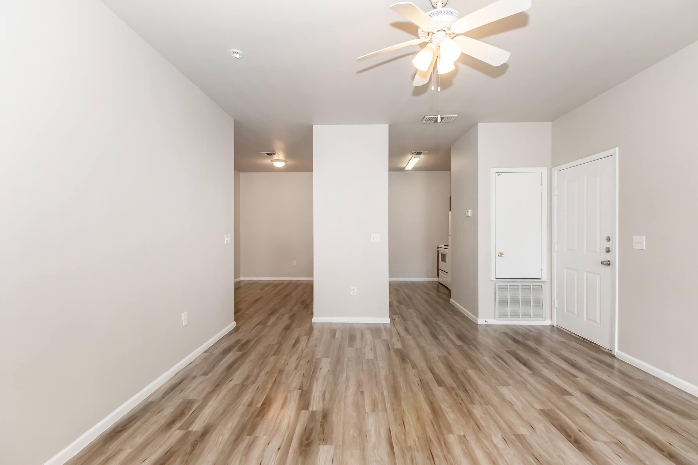Spacious interior of an apartment featuring light-colored walls, laminate flooring, and a ceiling fan. The layout includes a doorway on the right leading outside, a small hallway in the back, and an open living area, creating a bright and welcoming atmosphere.