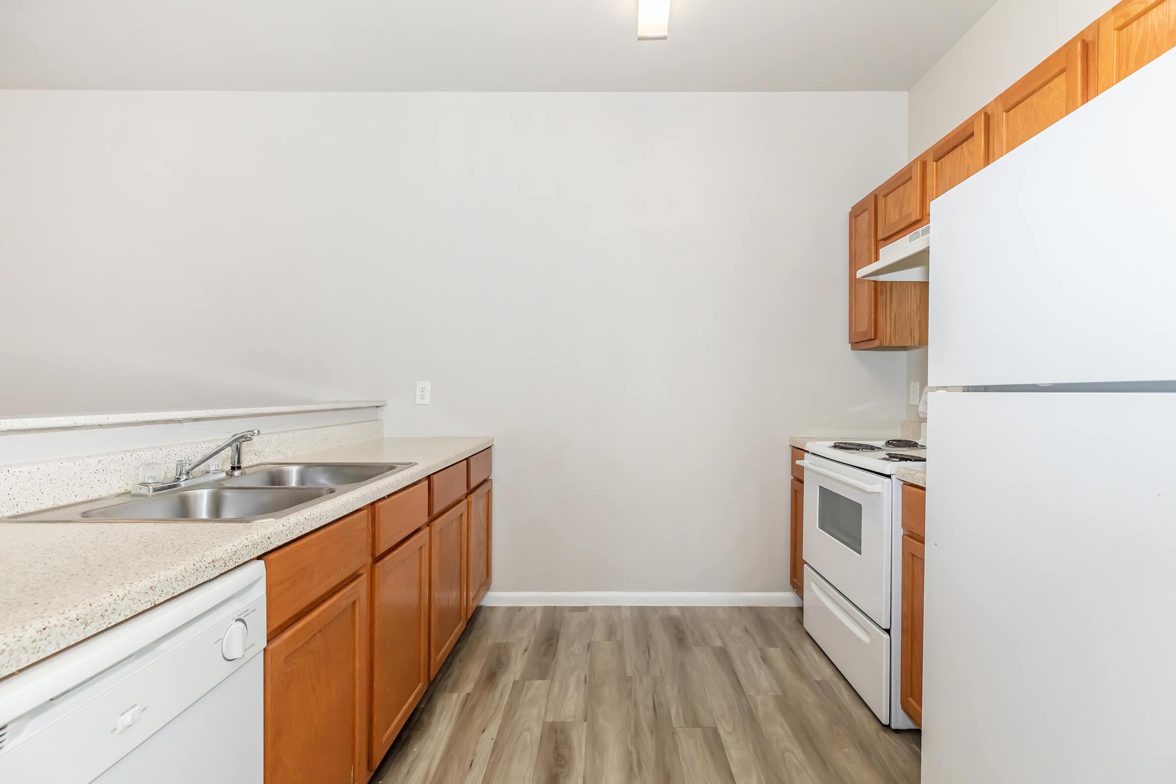 A modern kitchen featuring light-colored cabinetry, a double sink, a white stove, and a refrigerator. The floor is designed with wooden planks. The walls are a neutral shade, and the space is well-lit, creating an inviting atmosphere.