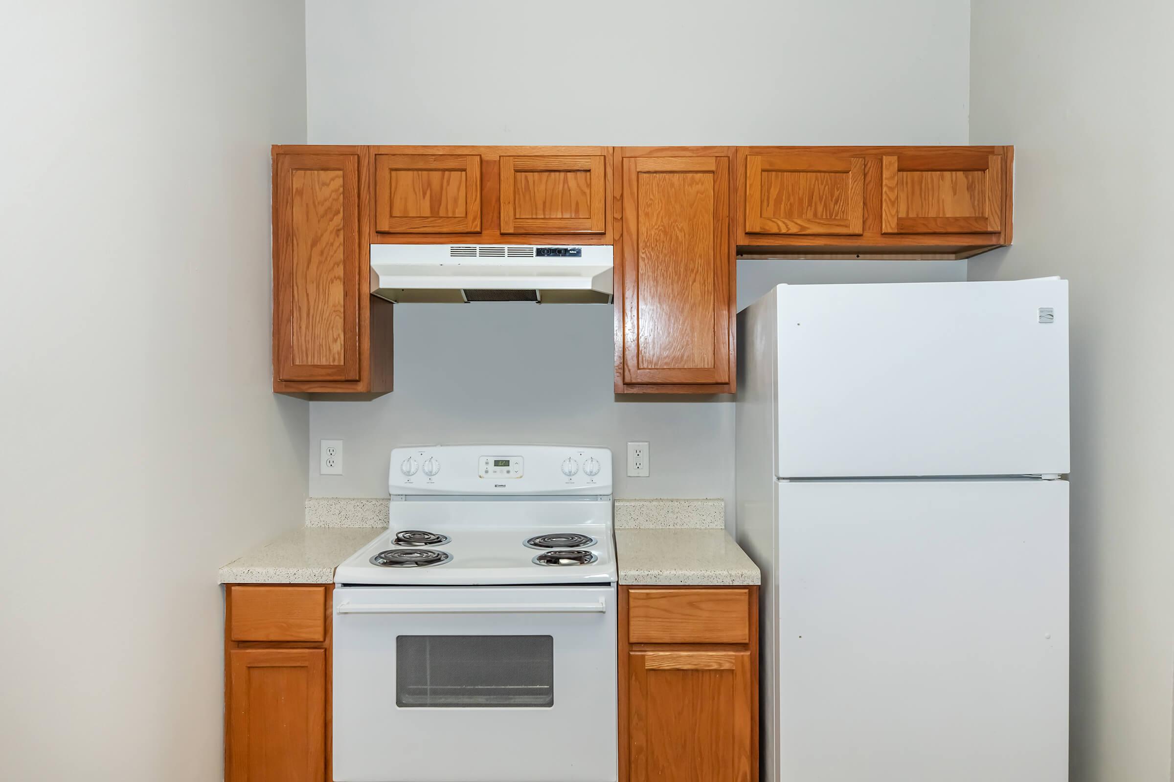 A compact kitchen featuring wooden cabinetry, a white refrigerator, a white stove with four burners, and a vent hood. The countertop has a light-colored finish, and the walls are painted a neutral tone, creating a simple and functional cooking space.