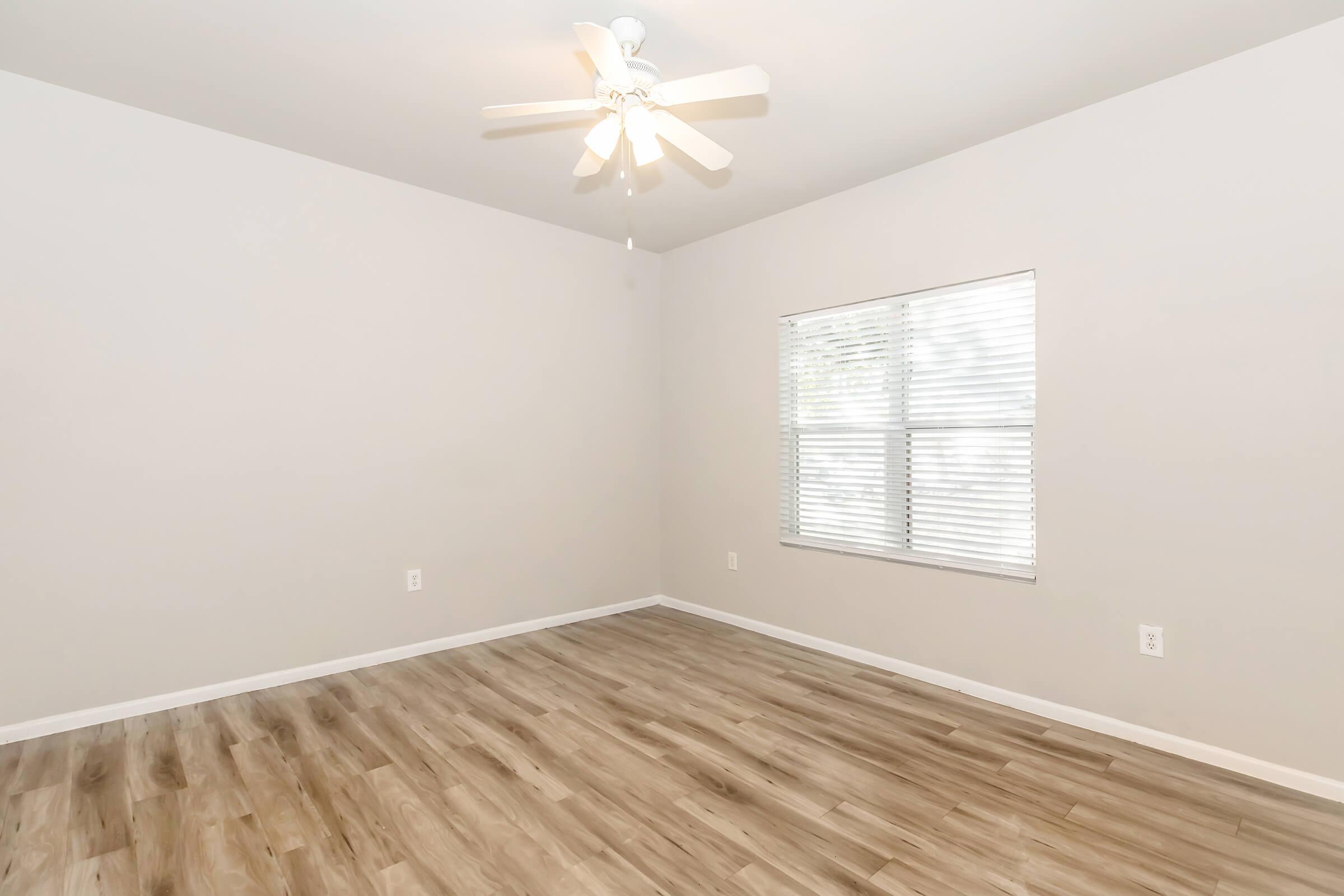 A spacious, empty room with light gray walls and a ceiling fan. The floor features light wood laminate. A window with white blinds allows natural light to enter, creating a bright atmosphere. The room is uncluttered, offering a fresh and clean appearance, ideal for personal decoration.