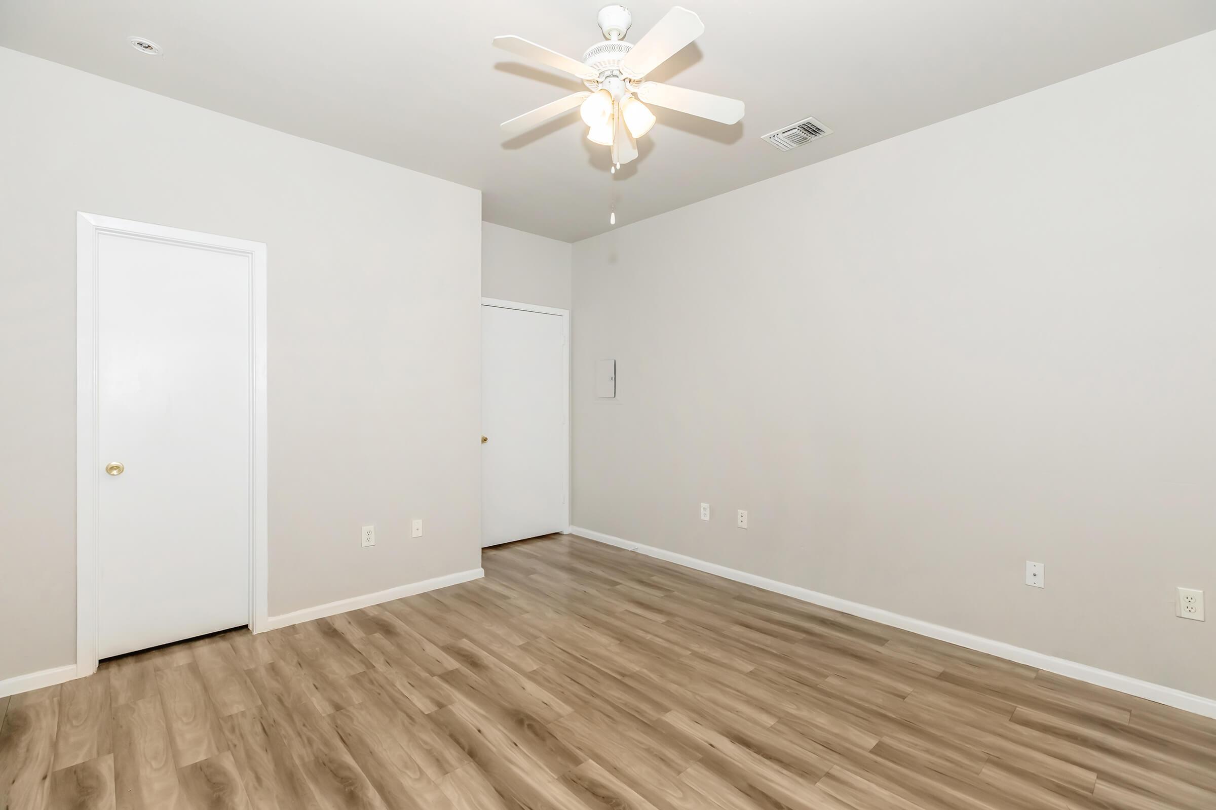 Empty room with light-colored walls and a ceiling fan. One door on the left and another on the right lead to separate spaces. The floor is light wood laminate, and there are no visible furnishings or decorations, creating an open and spacious feel.