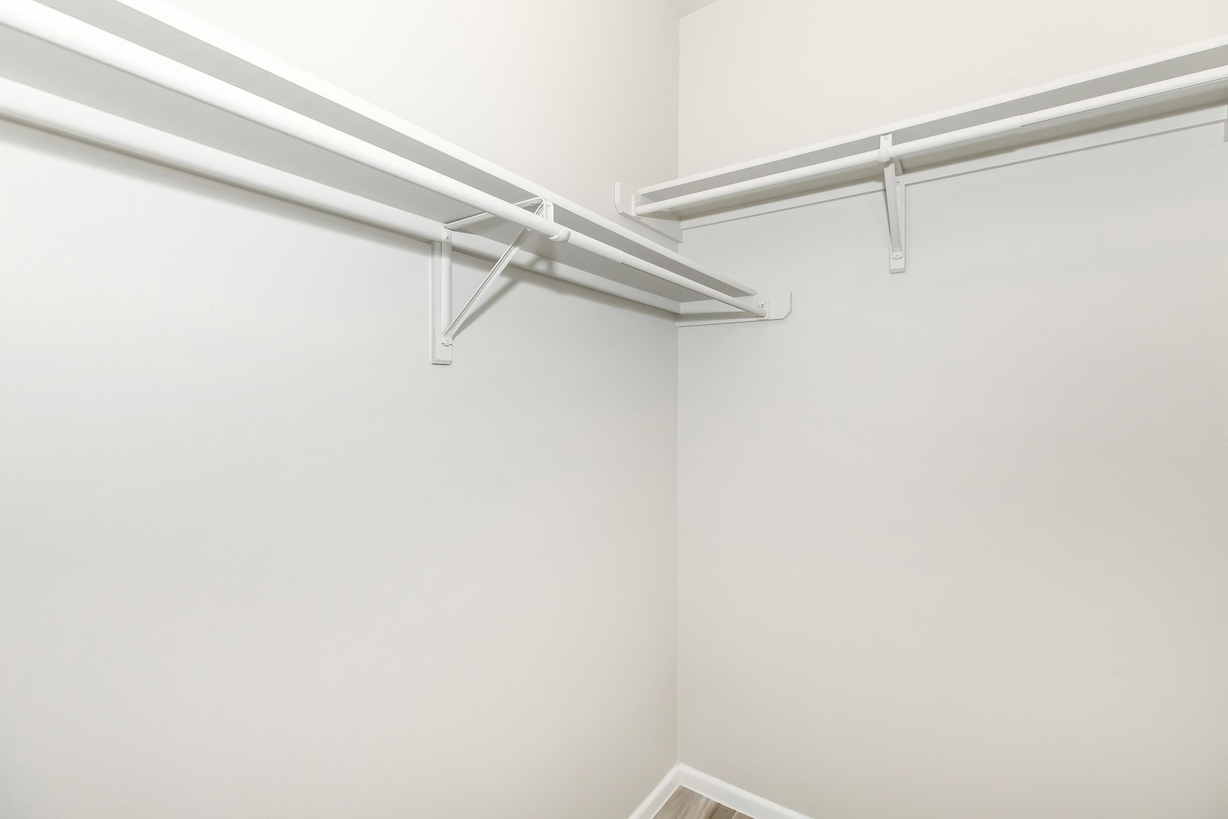 Empty closet space with two white shelving rods mounted on light gray walls. The floor is bare, and the overall design is minimalist, creating a clean and simple storage area.