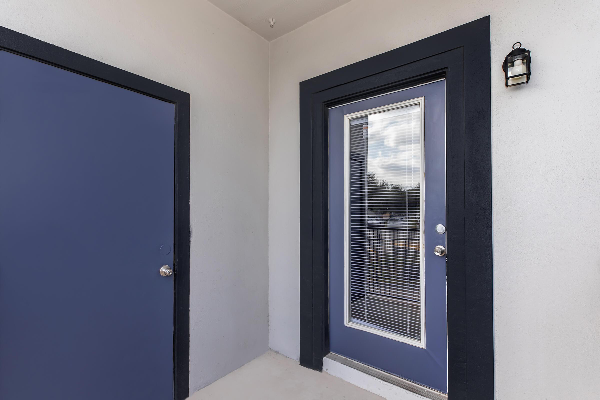 A modern entryway featuring a blue door with glass panels, flanked by a smaller blue door to the left. The area is well-lit with a light fixture mounted beside the door, and the walls are painted in a light color, creating a clean and inviting appearance.