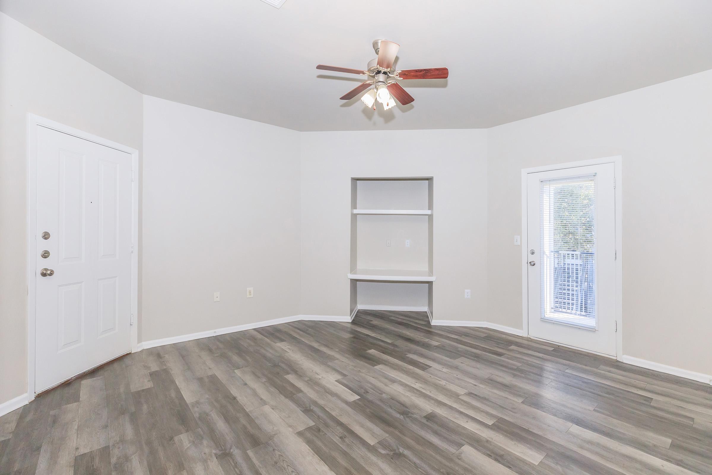 Empty room featuring light-colored walls, laminate flooring, a ceiling fan with wooden blades, and a small built-in shelf. There are two entrances: one door leading outside and another that appears to be the main entrance. Natural light enters through the door on the right.