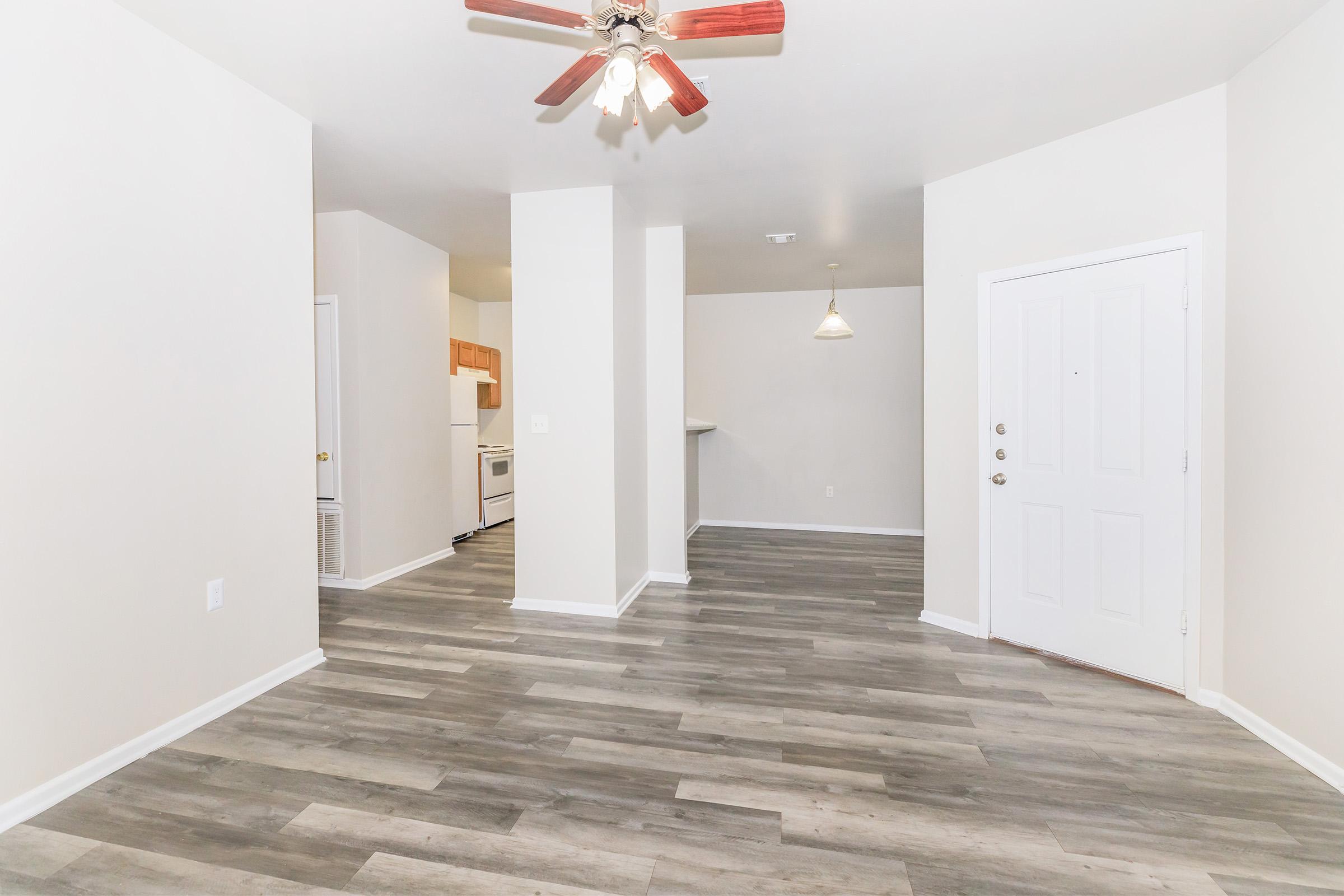 An empty living space with light-colored walls and a wood-style floor. Featuring a ceiling fan, an open layout leading to a kitchen area, and a front door. Natural light fills the room, enhancing the spacious feel.