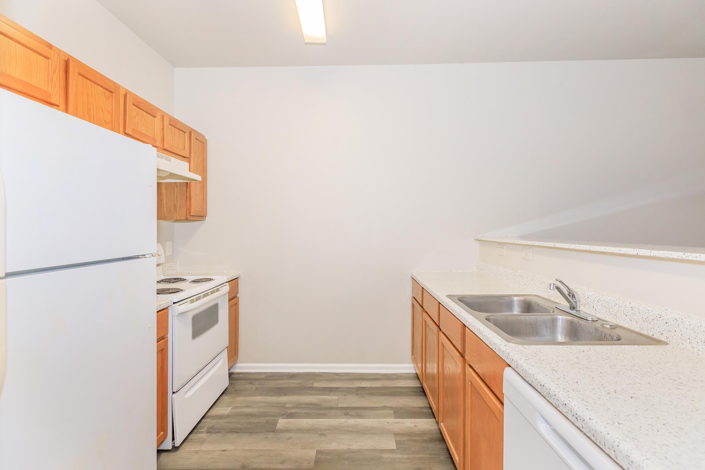 A modern kitchen featuring wooden cabinetry, a white refrigerator, an electric stove, and a double sink. The countertops are light-colored and speckled, with a neutral-toned floor, creating a clean and inviting cooking space. Natural light illuminates the room from a ceiling fixture.