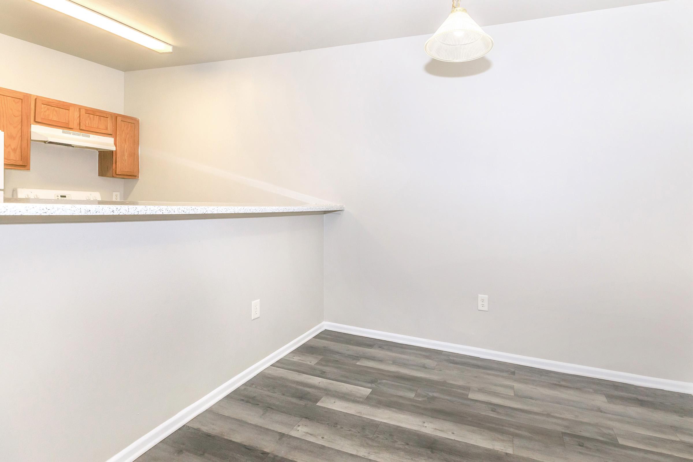 A bright, empty room with light gray walls and a wooden floor. In the background, there is a kitchen area with wooden cabinets and a countertop. A single light fixture hangs from the ceiling, illuminating the space.