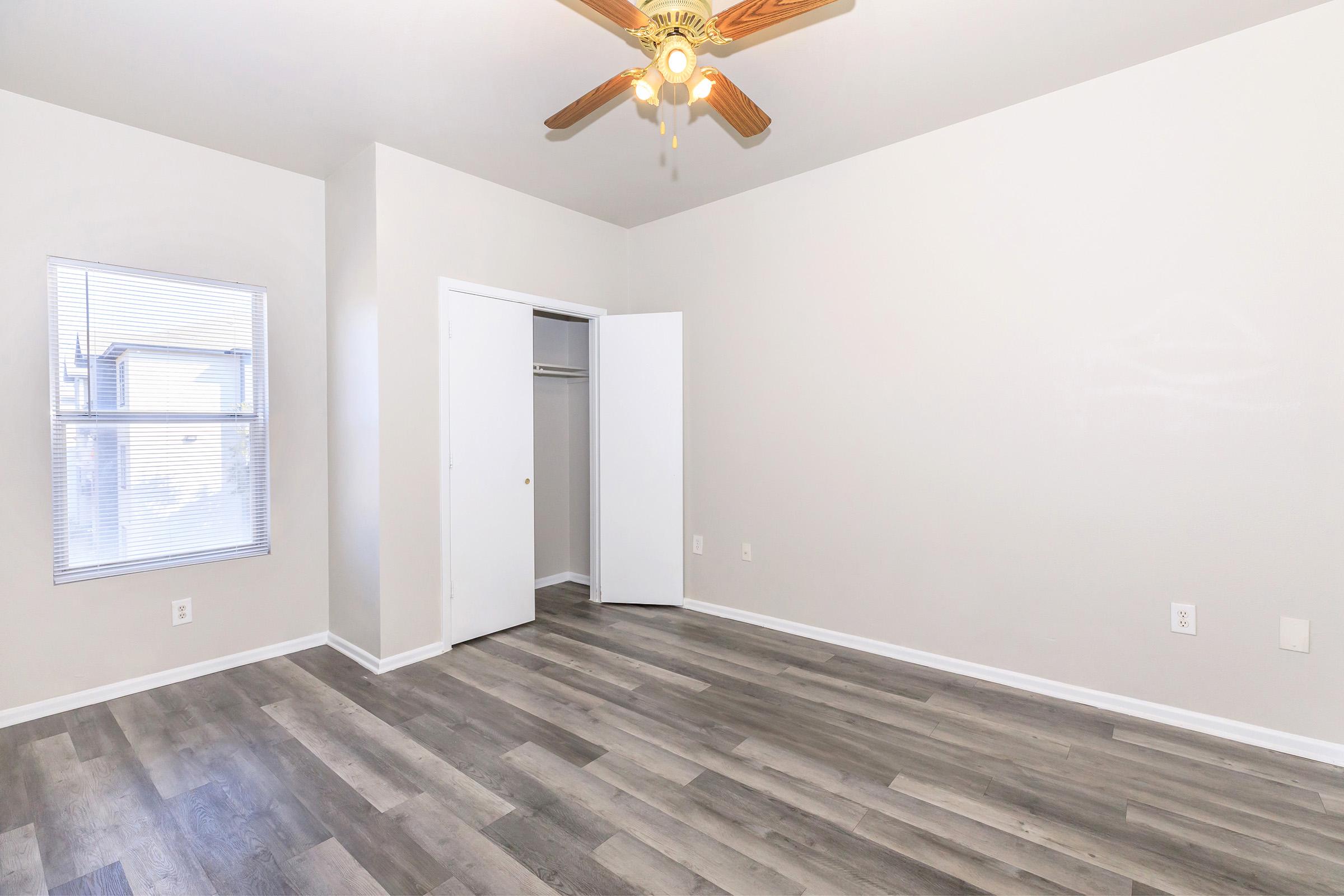 Empty room with light-colored walls and wood-like flooring. A ceiling fan with wooden blades is visible. There is a window on the left side, allowing natural light, and a closet with white doors is open on the right side. The overall space appears clean and minimalist.