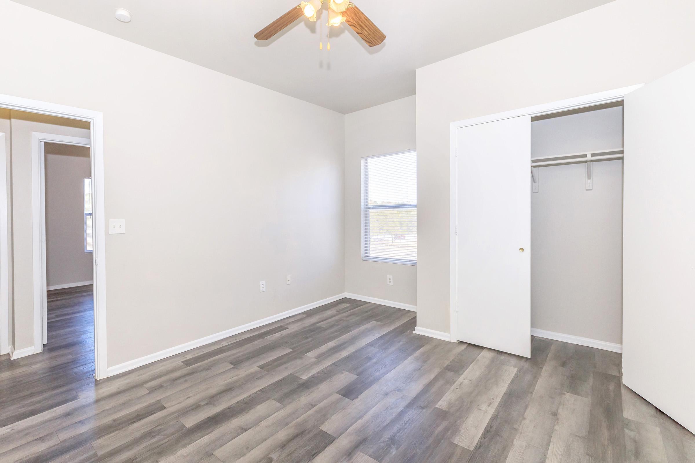 A bright, empty room with light gray walls and a ceiling fan. It features a window with blinds and a closet with white doors. The floor is made of light-colored laminate. The space appears clean and spacious, with an inviting atmosphere suitable for various uses.