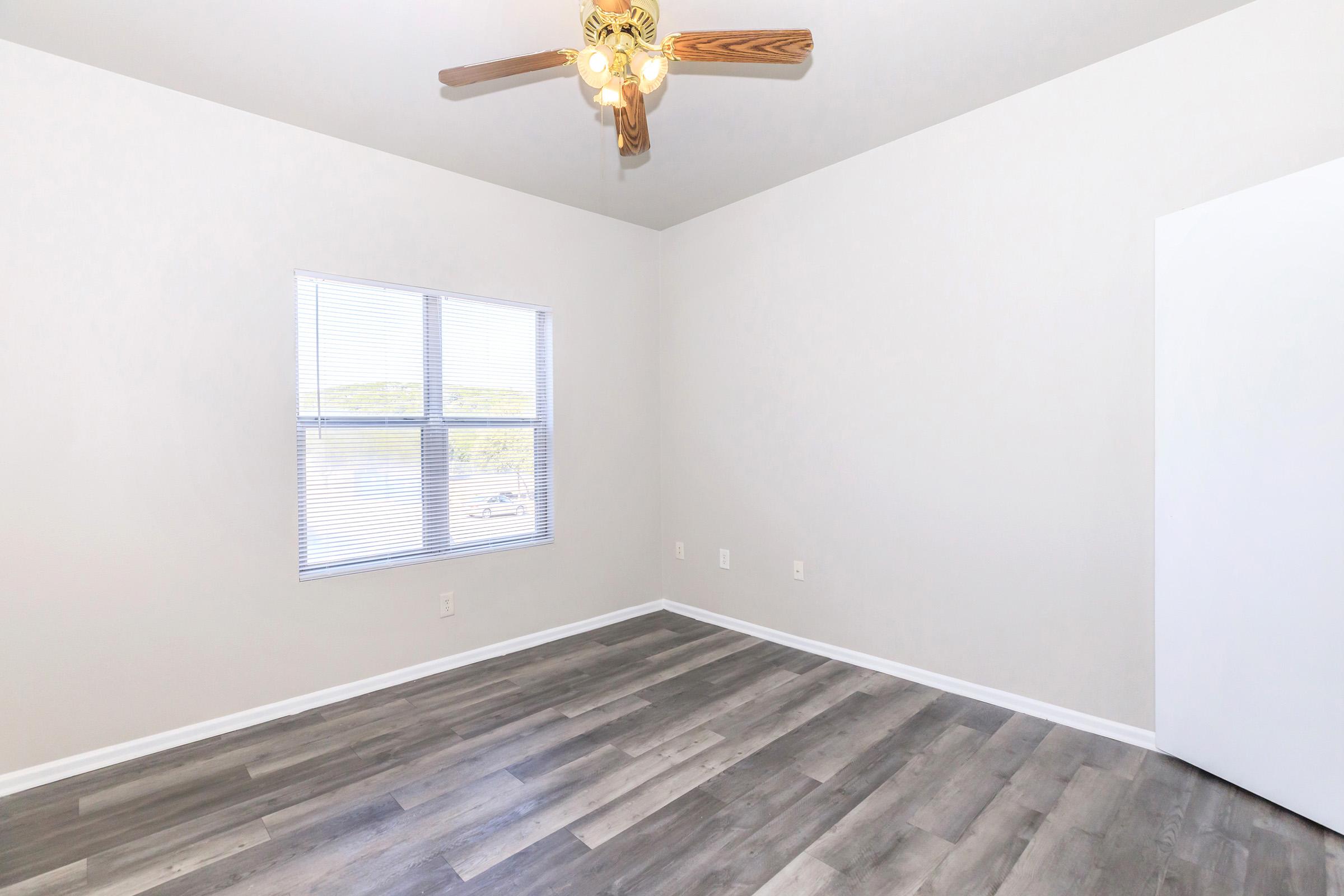 A bright and empty room featuring light-colored walls, a ceiling fan with wooden blades, and a large window with blinds. The floor is covered in gray laminate planks, creating a modern appearance. A white door is visible on the right side, and the overall ambiance is clean and inviting.