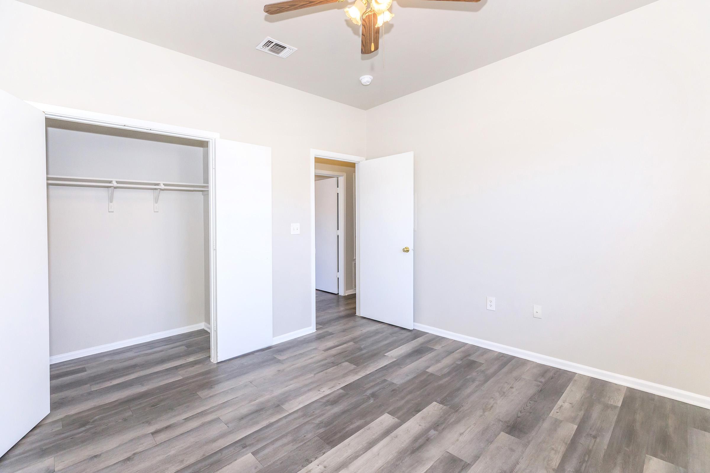 A brightly lit room featuring light-colored walls and laminate flooring. There are two open doors: one leading to a closet with hanging space, and the other possibly leading to a hallway. A ceiling fan is visible, adding a touch of comfort to the minimalist design.