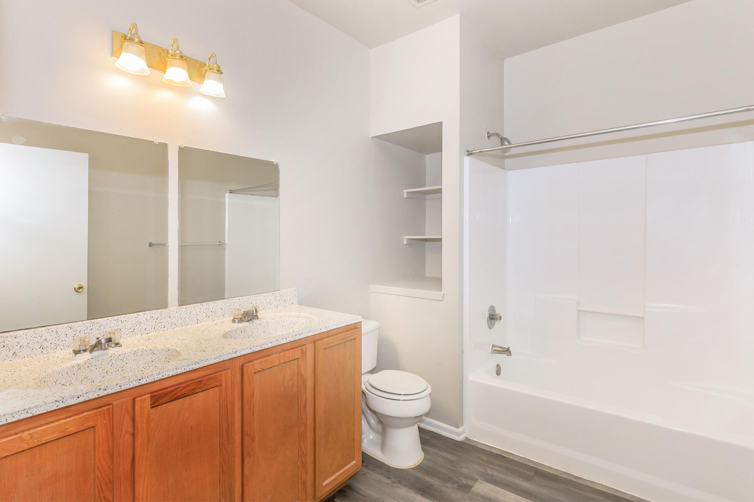A modern bathroom featuring a double sink vanity with granite countertops, two mirrors above the sinks, a white bathtub with shower, and a toilet. The walls are painted light, and there are built-in shelves beside the bathtub. The flooring is dark laminate.