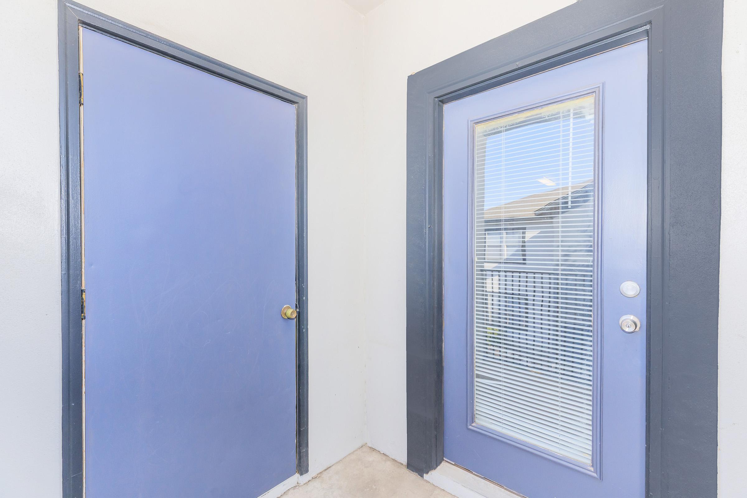 A hallway featuring two doors: one solid blue without windows and another blue with a glass panel that has blinds. The walls are light-colored and the flooring appears to be bare concrete. Natural light is coming from the glass door, illuminating the space.
