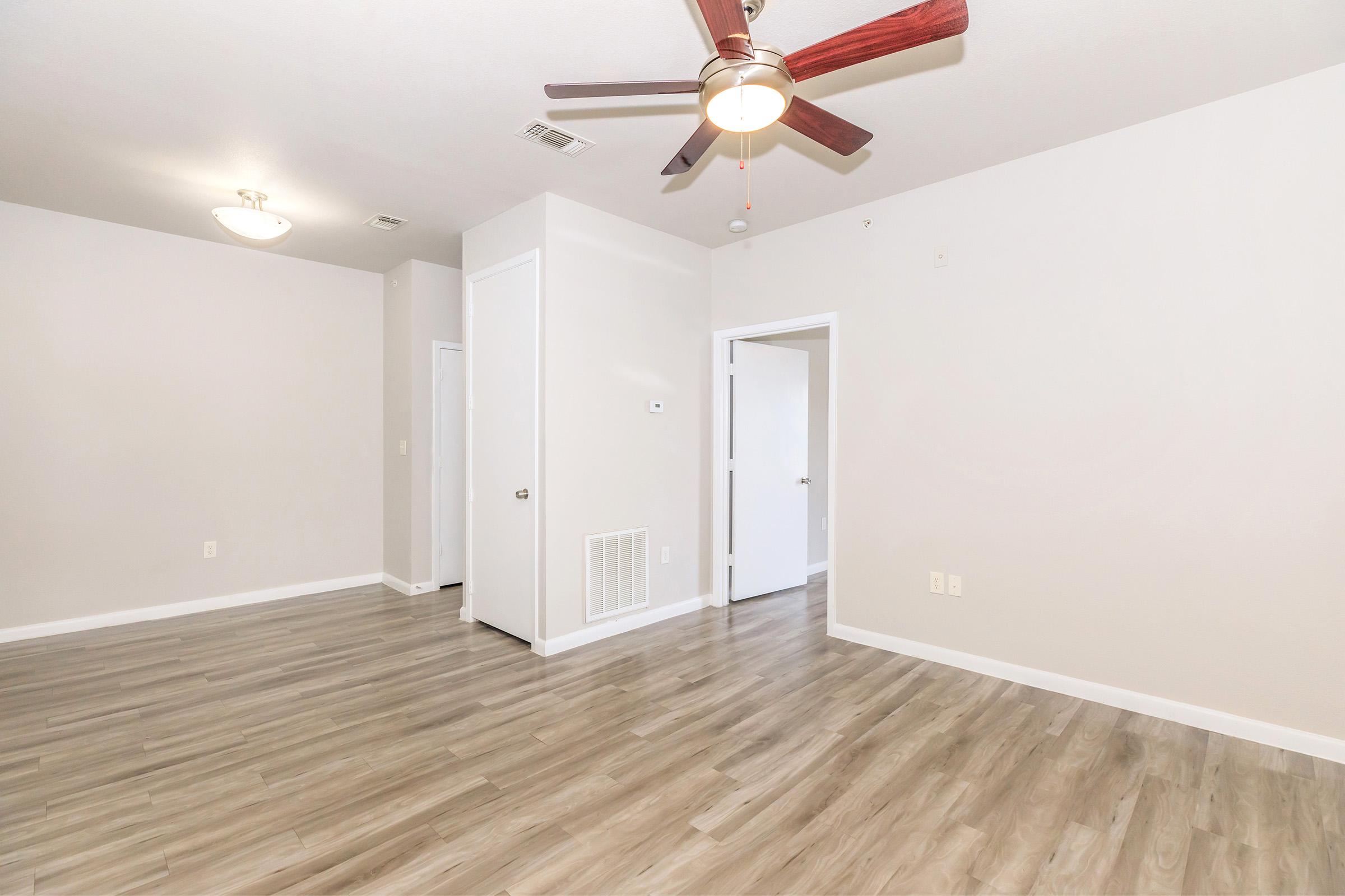 A spacious room with light-colored walls and wooden flooring. A ceiling fan with wooden blades hangs overhead. There are two doors leading to other spaces, and a wall vent is visible. Natural light brightens the area, creating an inviting environment.