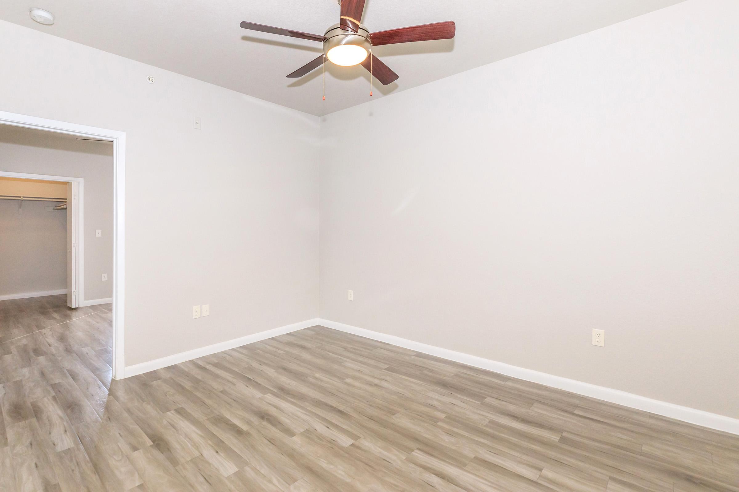 A spacious, empty room with light gray walls and a ceiling fan with wooden blades. The floor is covered in light brown laminate. There is an open doorway on the left leading to a closet, and the overall atmosphere is bright and clean with plenty of natural light.