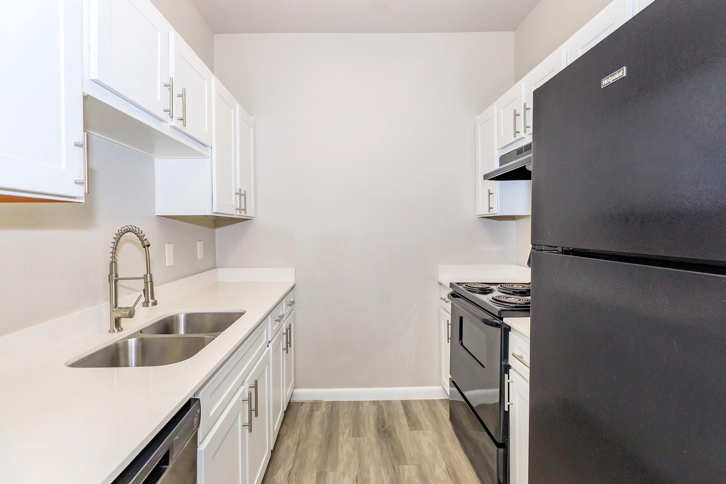 A modern kitchen featuring white cabinets, a double sink, a black refrigerator, and a black stove with an oven. The space has light-colored walls and a clean, minimalist design with light wood-like flooring.