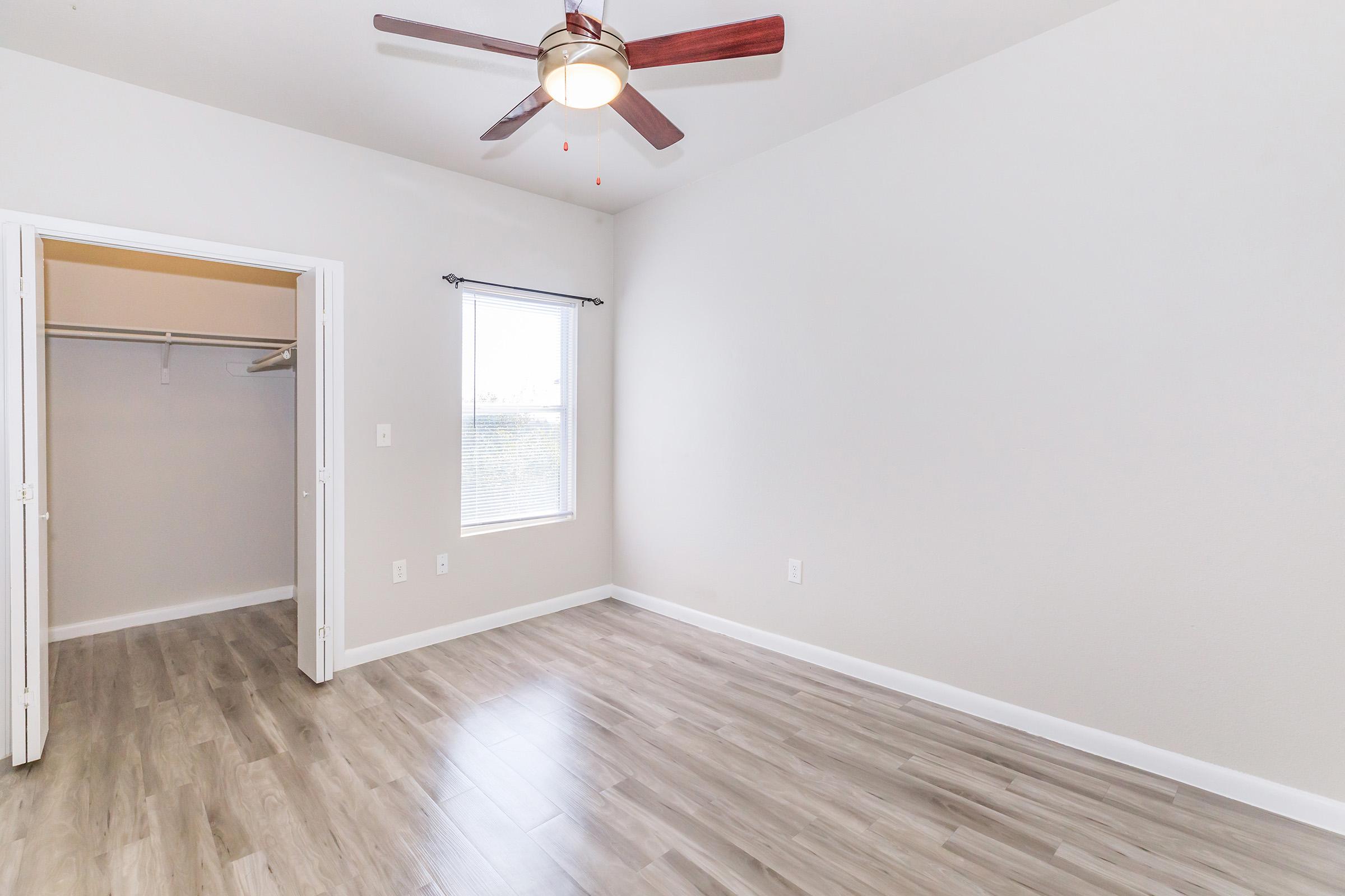 A spacious, empty room with light gray walls and wood-look laminate flooring. It features a ceiling fan with brown blades and a window letting in natural light. A closet with double doors is visible on the left side, emphasizing the room's openness and potential for personalization.