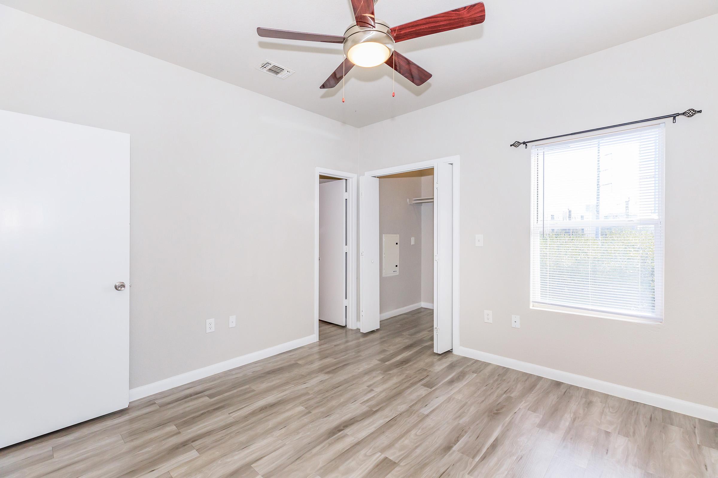 A bright, empty room featuring light wood-like flooring, a ceiling fan with wooden blades, and neutral-colored walls. It has a window with blinds allowing natural light, and two doorways leading to other areas. The room conveys a spacious and clean feel, suitable for various furnishings.