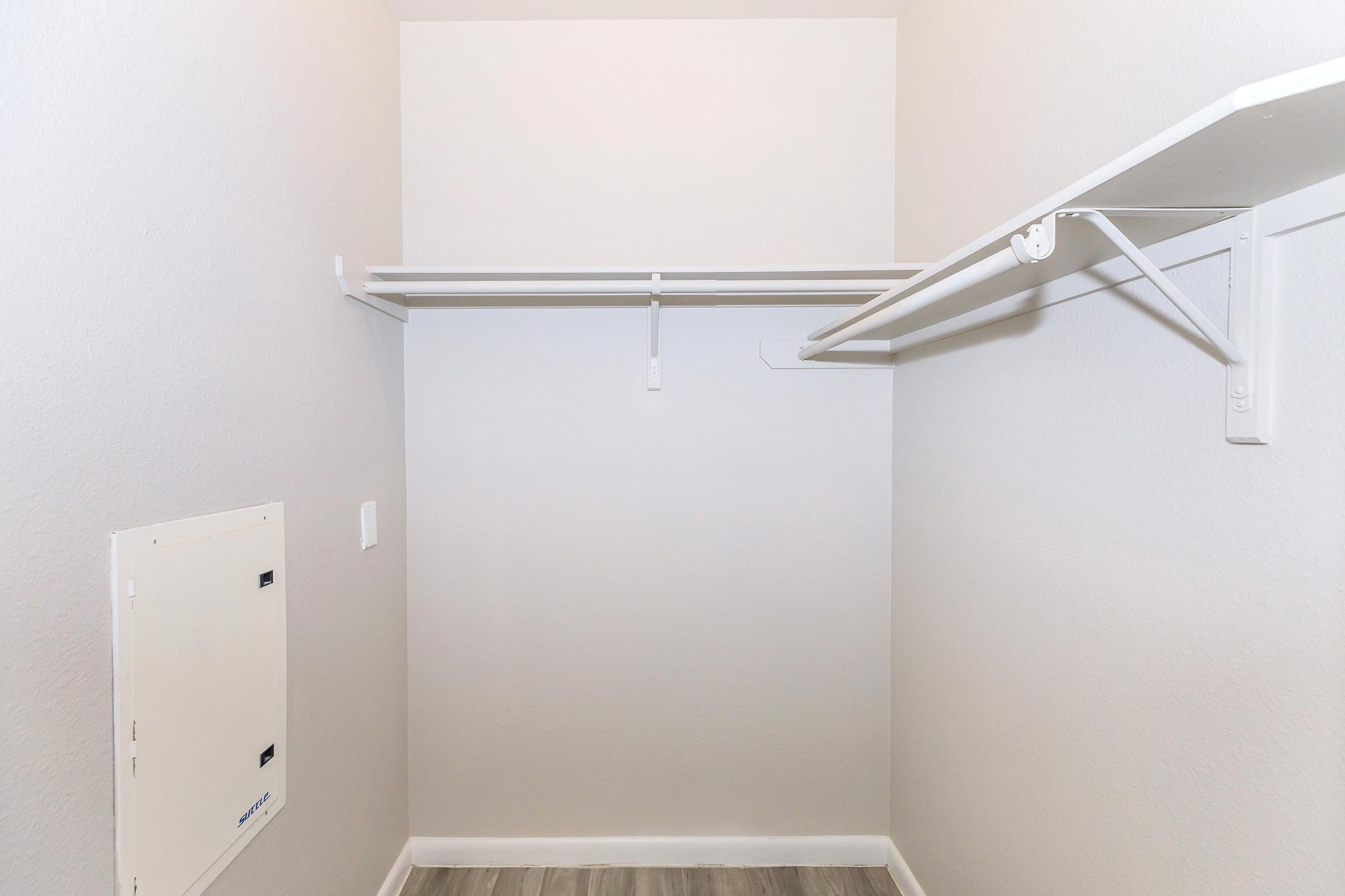 Empty closet with a light gray wall, featuring two white shelving units for hanging clothes. The floor has a light wood-like finish, and there's a small access panel on the left wall. The overall space is clean and minimalistic.