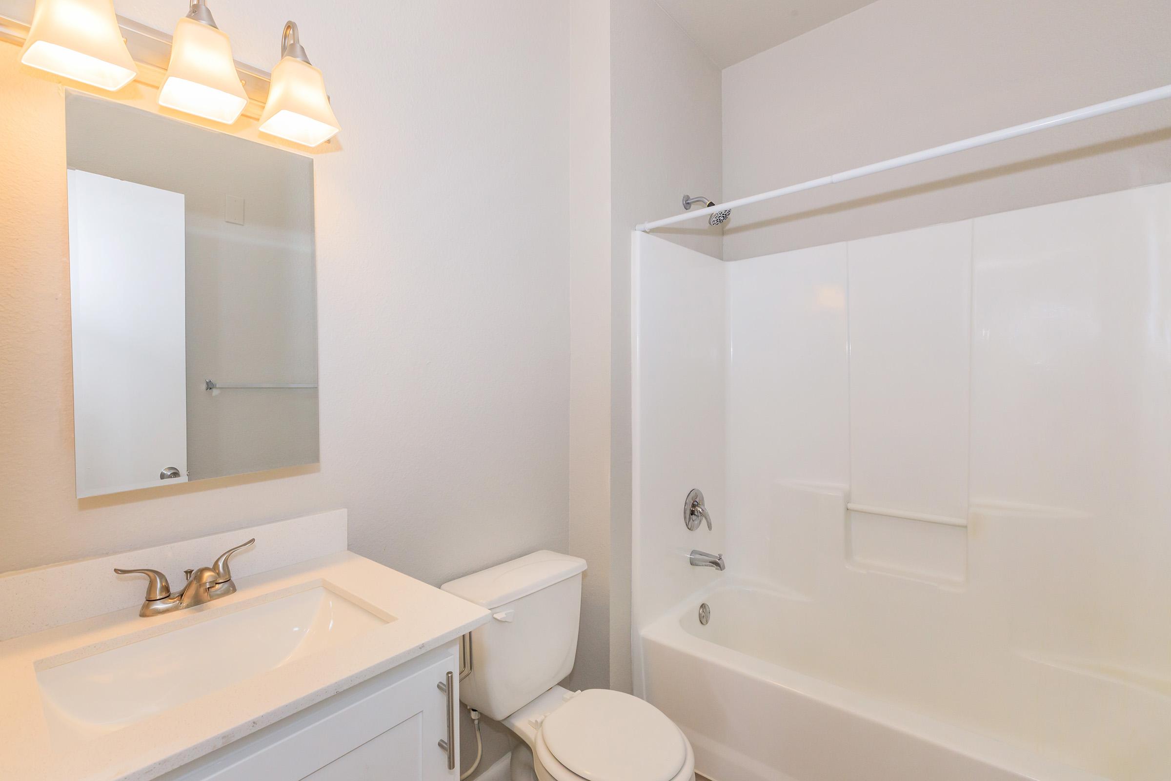 A clean and modern bathroom featuring a white bathtub with a shower, a white toilet, and a vanity with a sink. Three light fixtures are mounted above the mirror, and the walls are painted in a light, neutral color, creating a bright and spacious feel.