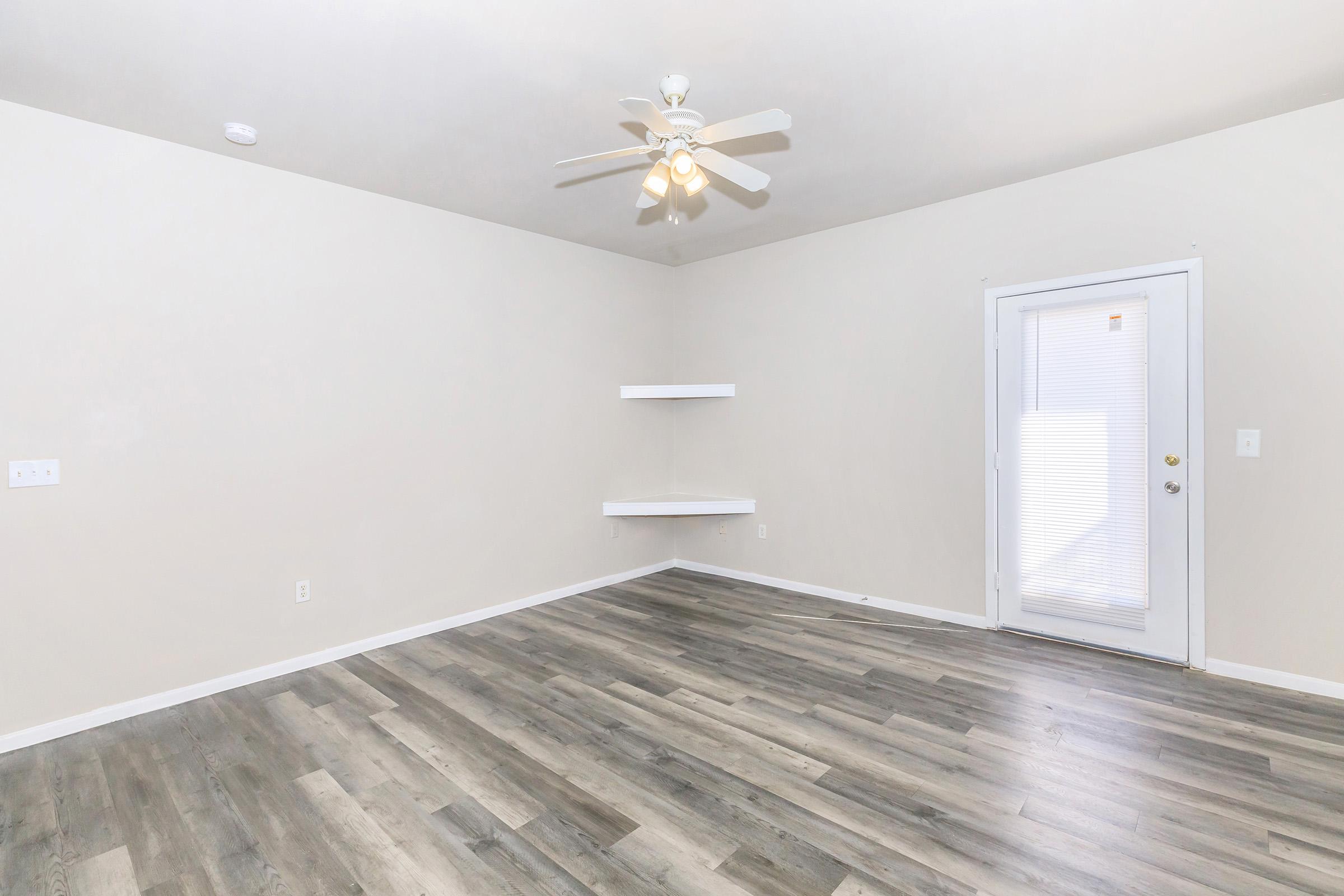 A spacious, empty room with light-colored walls and a ceiling fan. The floor features wooden planks, and there is a small white shelf on one wall. A glass door leads outside, letting in natural light. The overall atmosphere is bright and minimalistic, suitable for various interior design options.