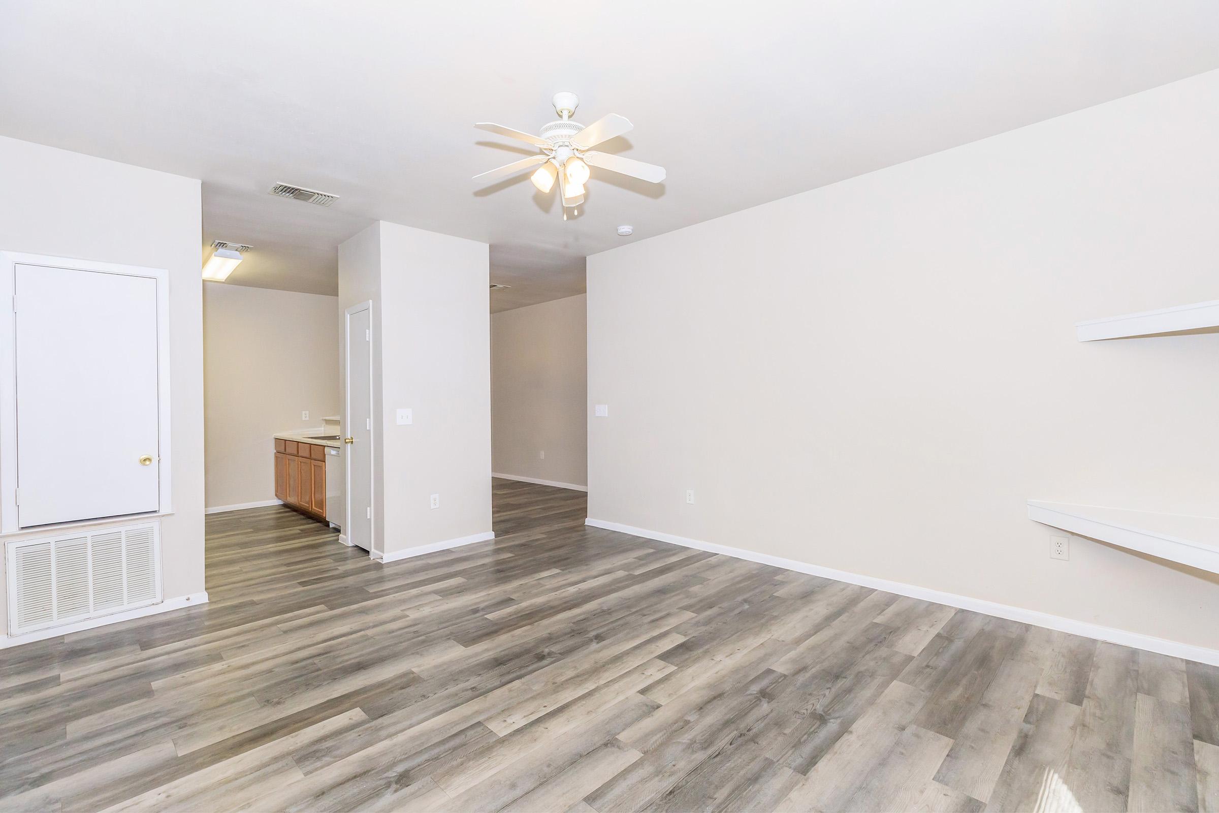 Spacious, light-filled interior of an empty room featuring a ceiling fan, light-colored walls, and modern laminate flooring. A doorway leads to a kitchen area with wooden cabinetry, and a small shelf is mounted on the wall, indicating a living space ready for decoration and personalization.