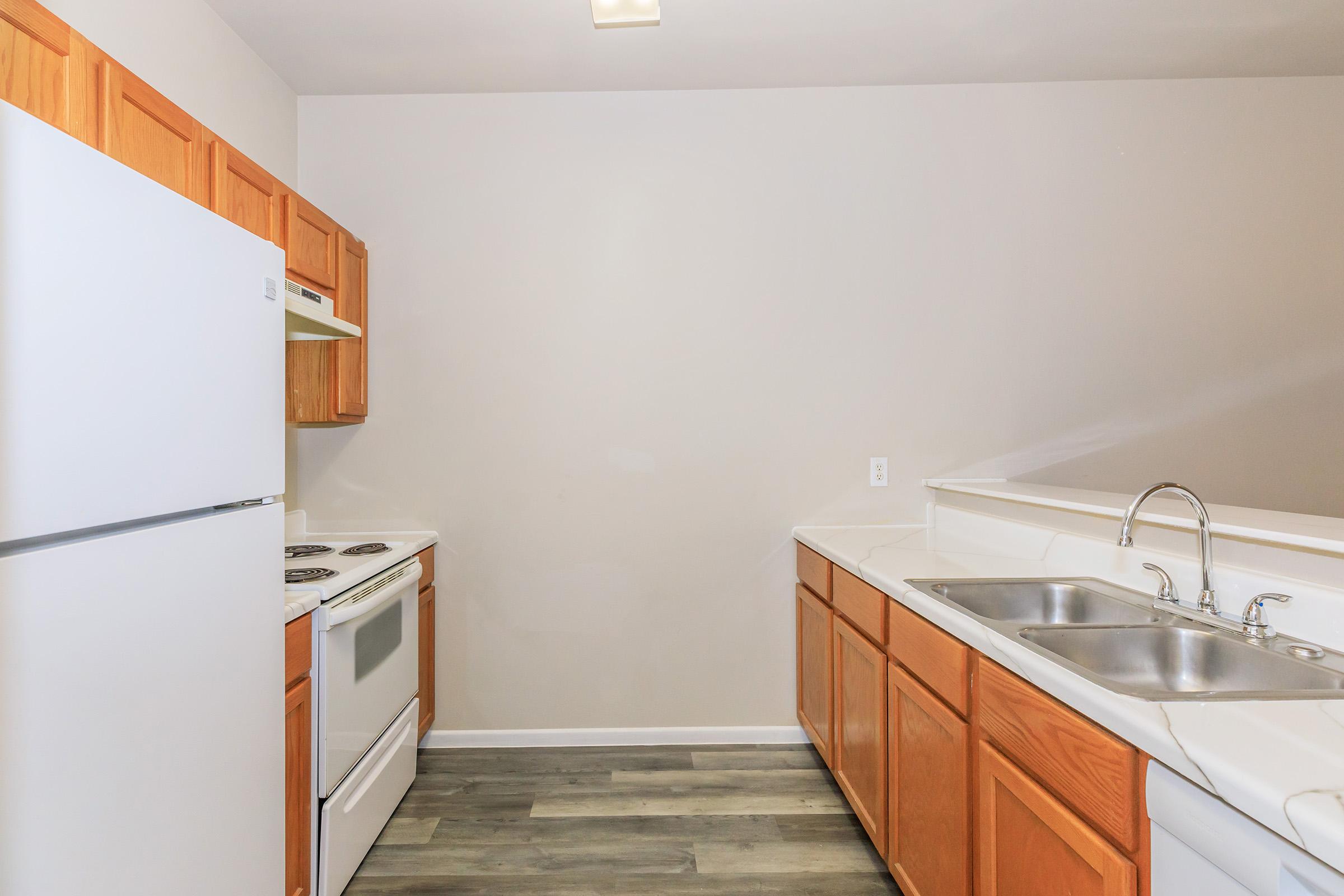 A modern kitchen featuring wooden cabinets, a white refrigerator, an oven, and a two-basin sink. The walls are painted light gray, and the flooring is a light wood pattern, creating a clean and inviting space.