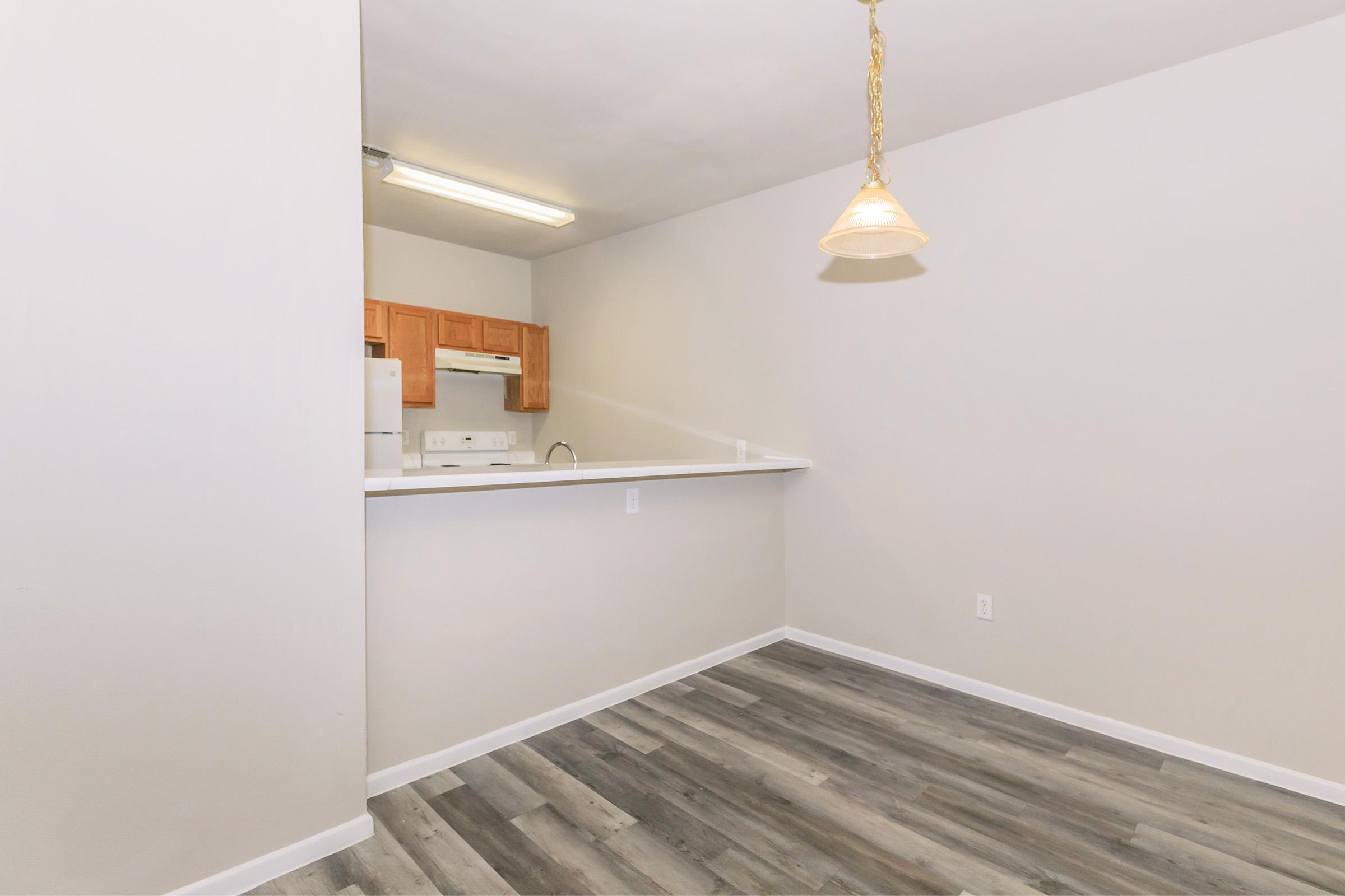 A light-filled kitchen area featuring beige walls and gray wood flooring. The space includes a bar counter that opens to the kitchen, where wooden cabinets and white appliances are visible. A pendant light hangs from the ceiling, adding to the modern decor. The overall ambiance is clean and inviting.