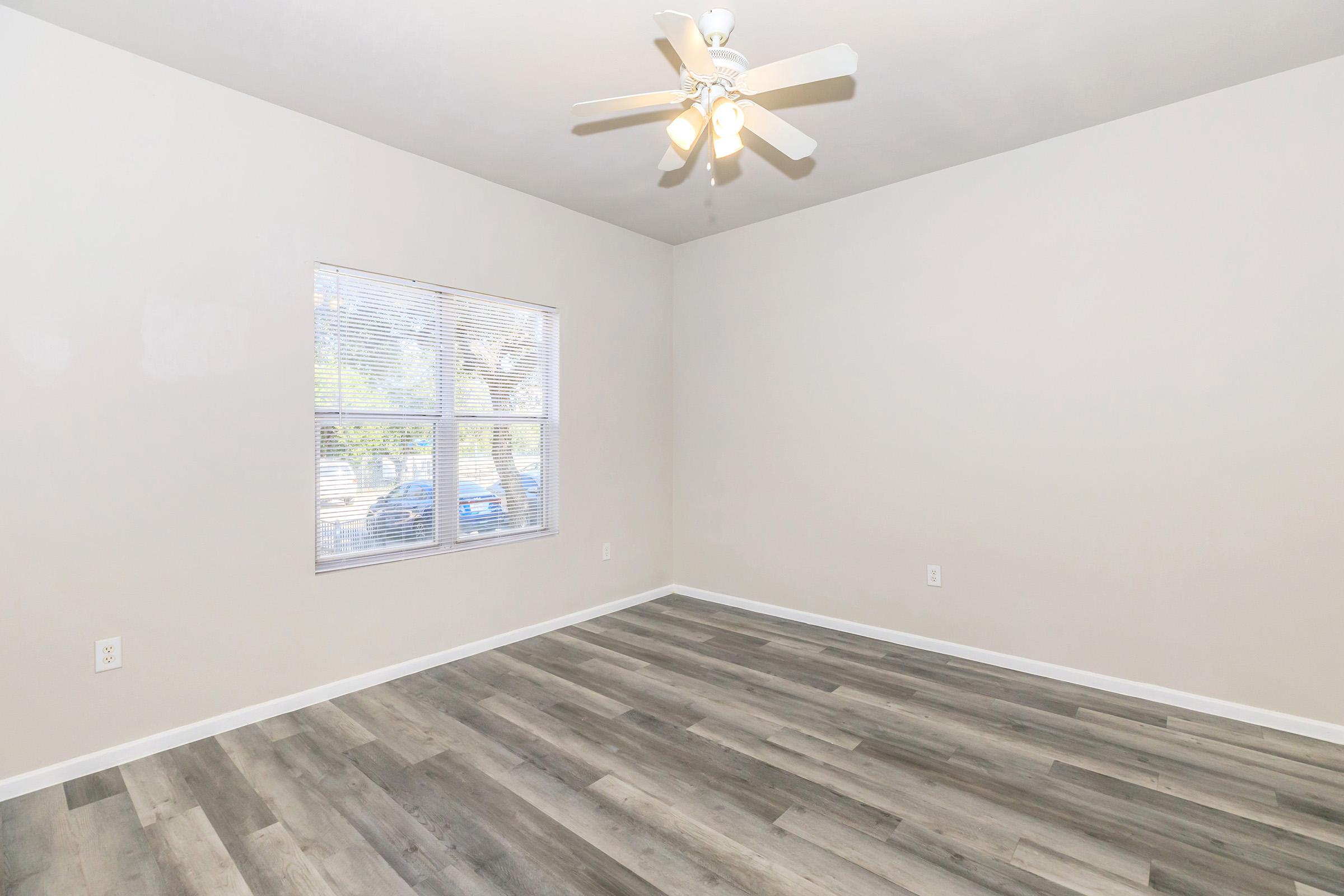 Empty room with light gray walls and a ceiling fan. A large window allows natural light to enter, showing a view of parked cars outside. The floor features a modern, wood-like design. The space is clean and minimal, suitable for various uses or decoration.