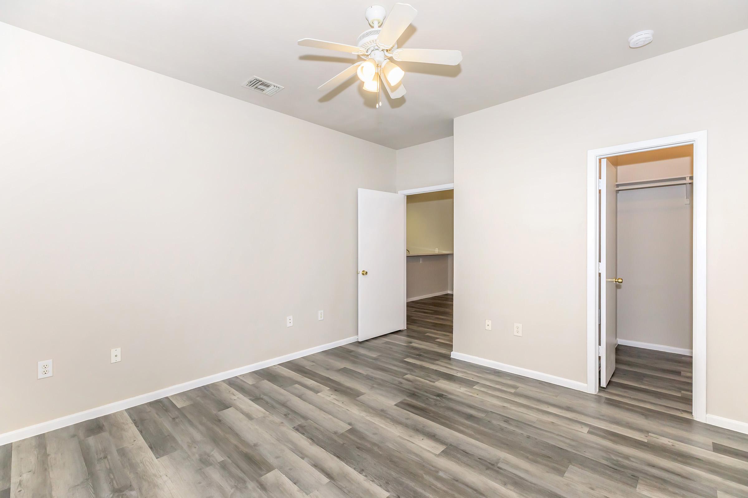 A spacious, unfurnished room with light-colored walls and a wooden floor. A ceiling fan is mounted above, and there is an open door on one side leading to a closet. The room has a neutral ambiance, ideal for customization.
