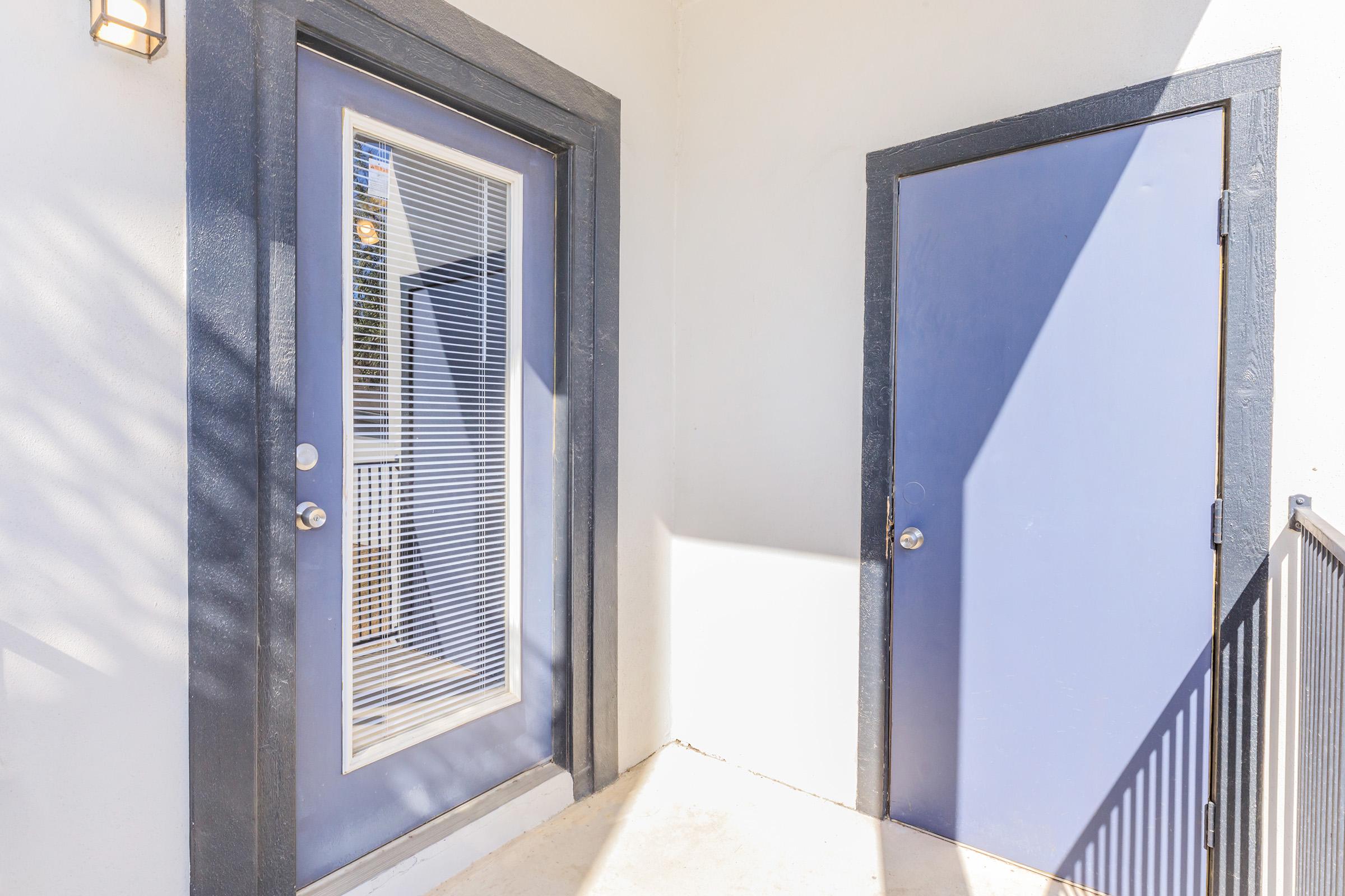 A well-lit entrance area with two doors. The door on the left has a window with blinds, while the door on the right is solid and painted blue. The walls are light-colored, and there is a railing visible in the bottom right corner, suggesting an outdoor space. Shadow patterns add visual interest to the scene.